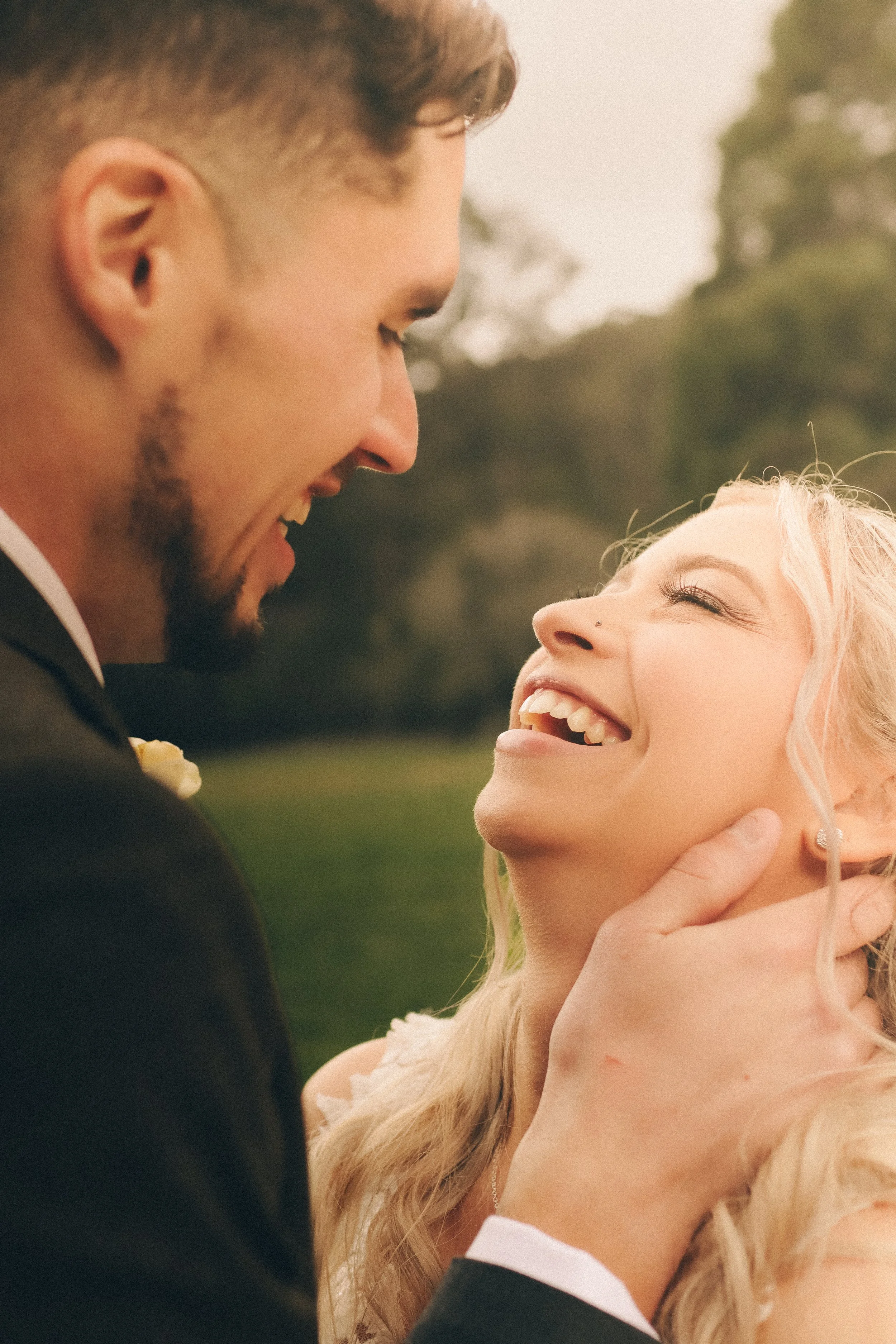 A man and woman smiling, embracing outdoors, the man holding the woman's face gently.