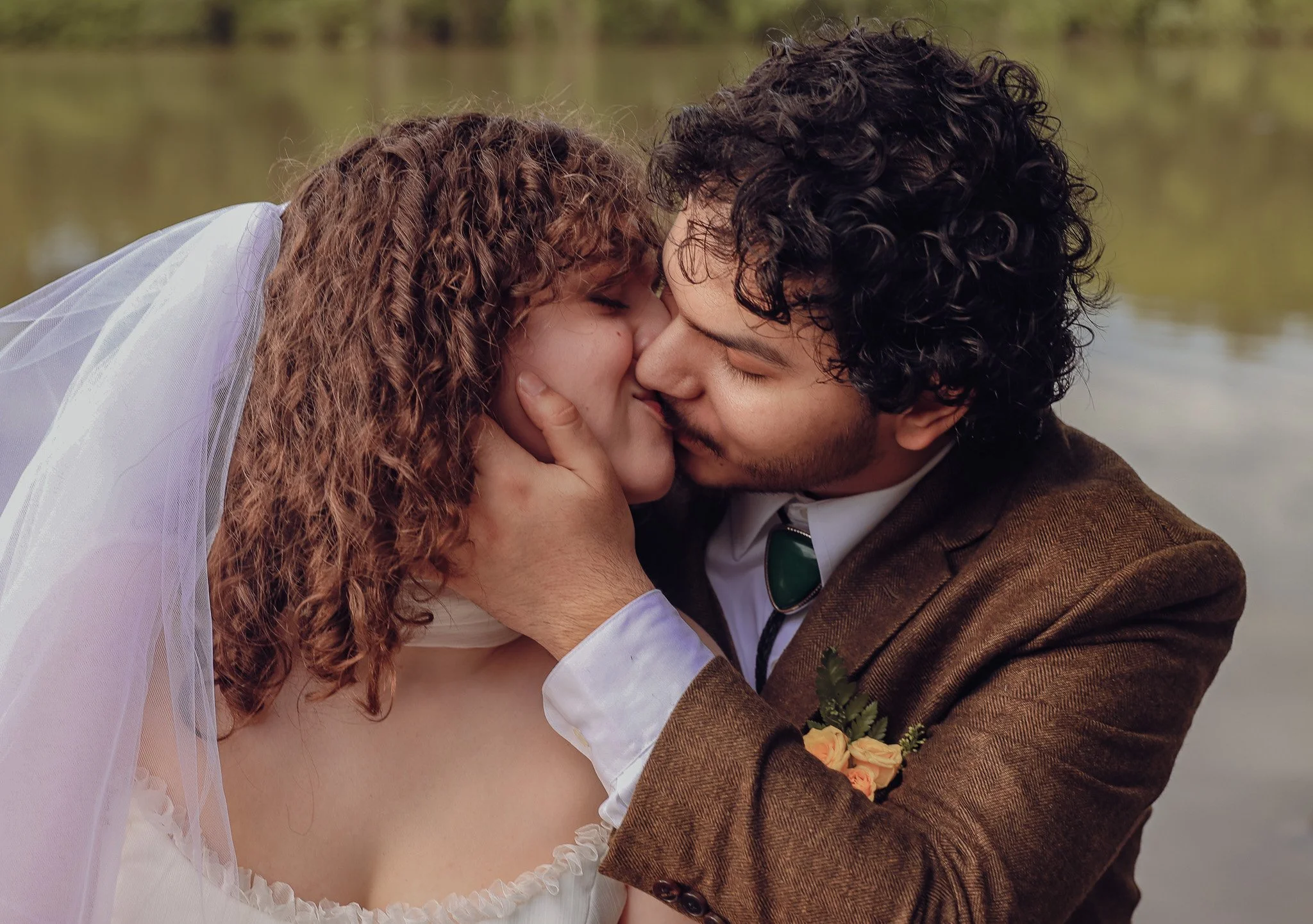 A couple sharing a kiss outdoors near a body of water, with the woman wearing a white veil and dress, and the man dressed in a brown suit with a boutonnière.