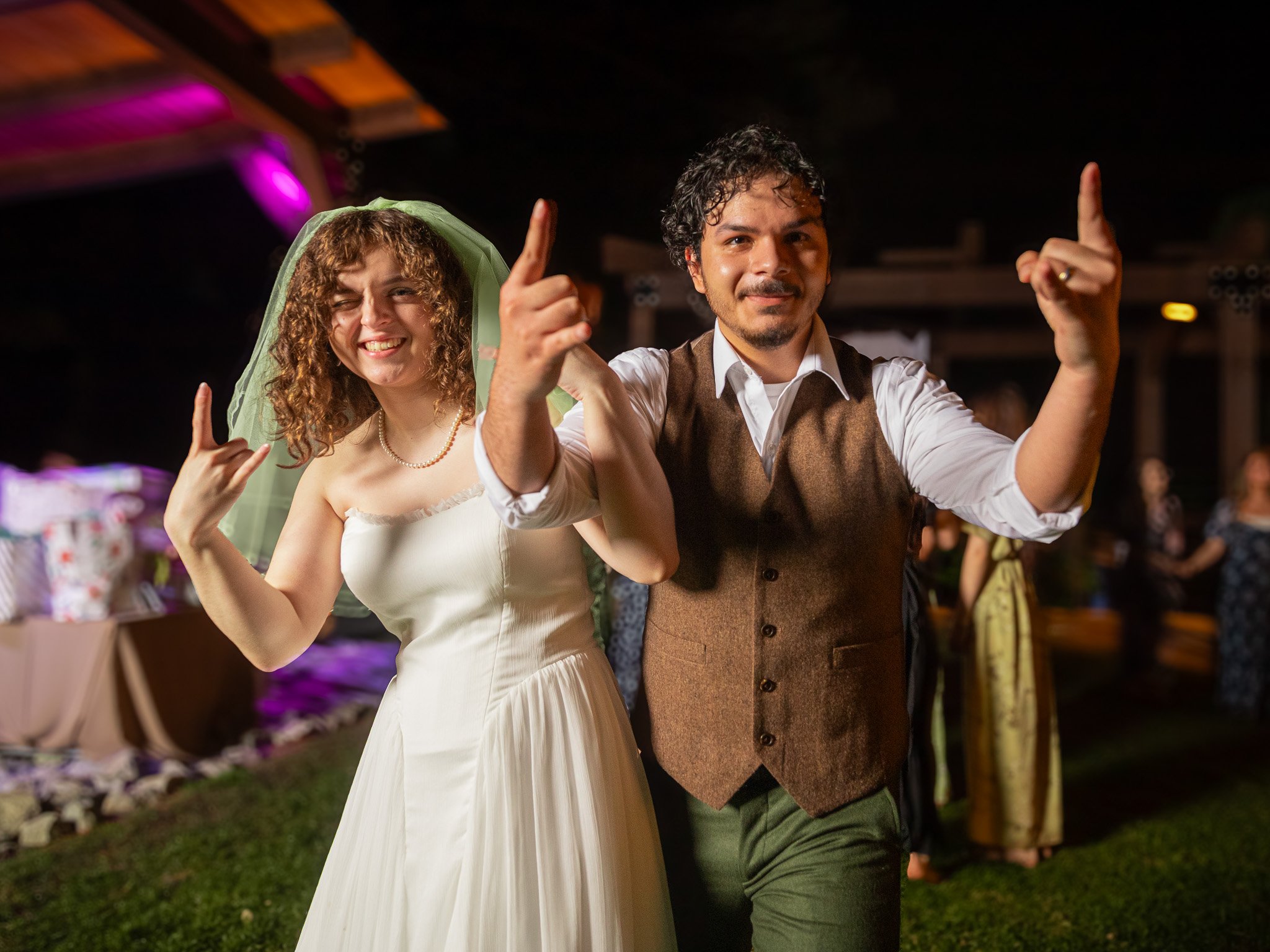 A smiling couple dancing at a wedding reception, the woman in a white wedding dress with a green veil, making rock on signs with her hands, and the man in a vest and white shirt, also making a rock on sign with his hands, in a dimly lit venue with ot