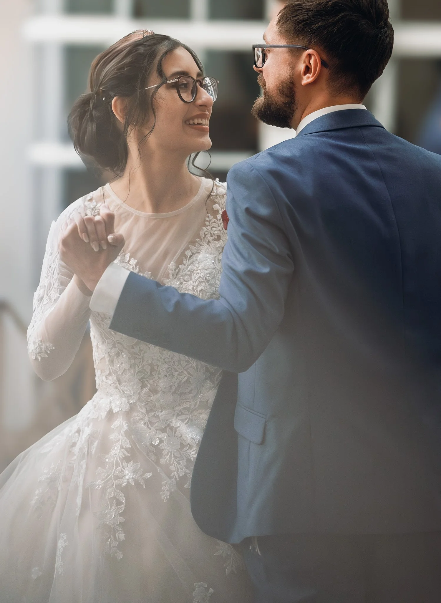 A bride and groom dancing together at their wedding, holding hands and smiling, indoors with blurred background.