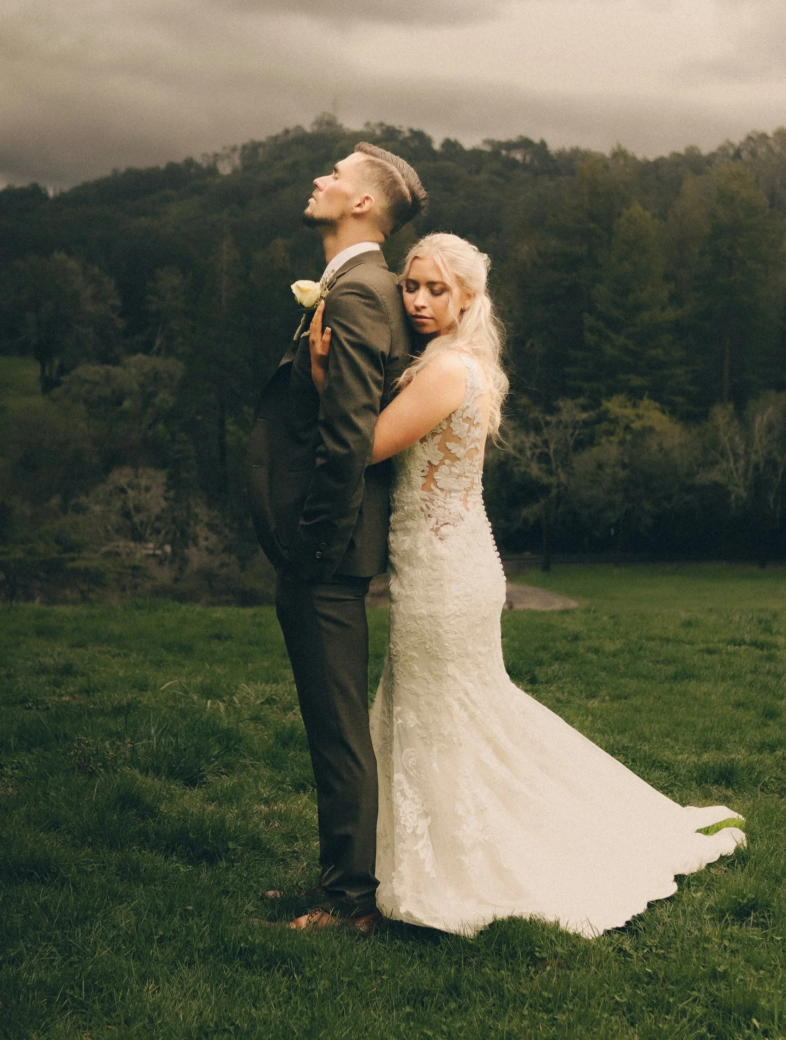A bride and groom standing outdoors on a grassy field with a backdrop of trees and hills, embracing each other during sunset or overcast weather.