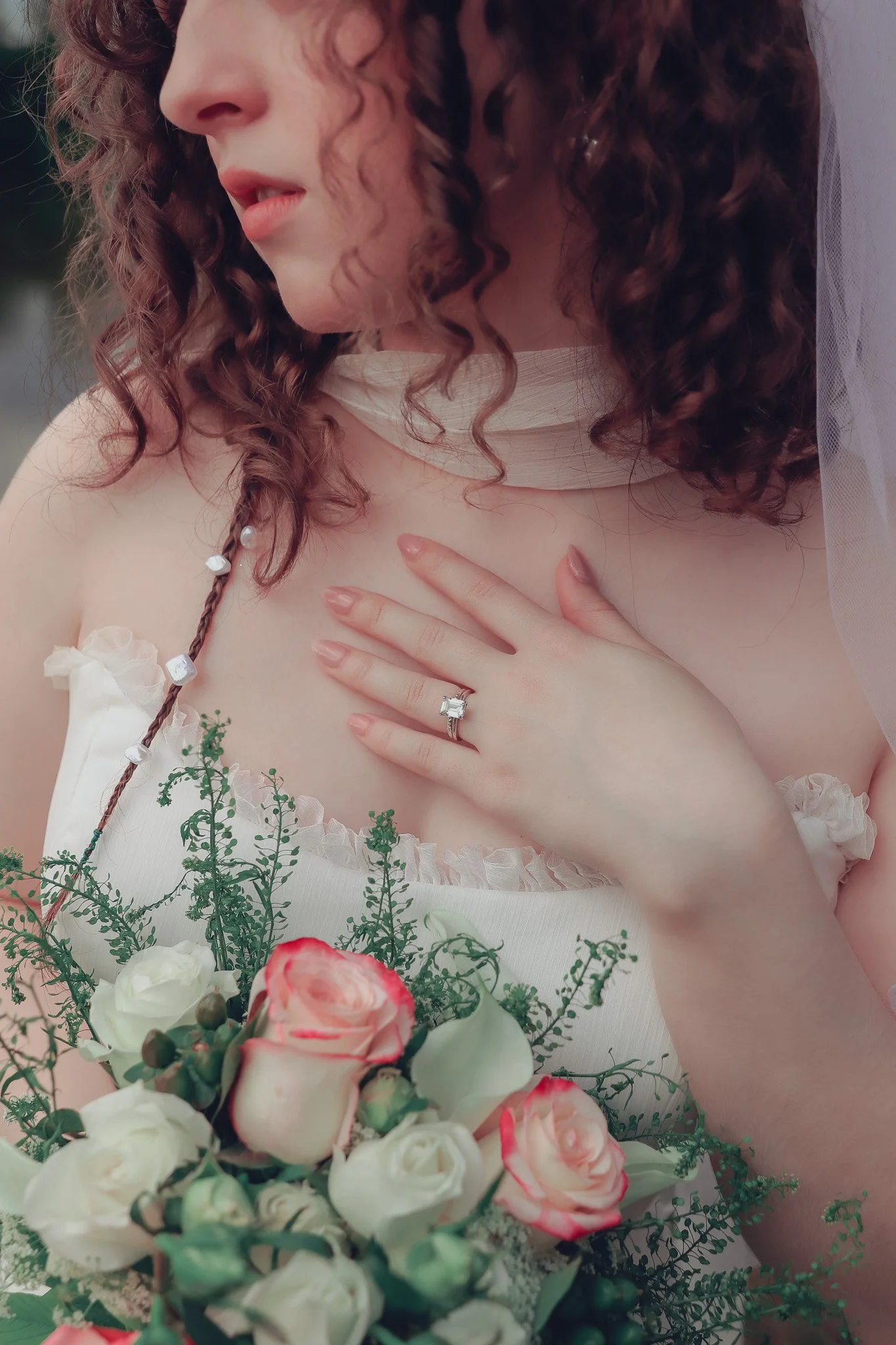 Close-up of a woman with curly hair wearing a wedding dress and veil, holding a bouquet of white and pink roses, with her hand over her chest showcasing a diamond engagement ring.