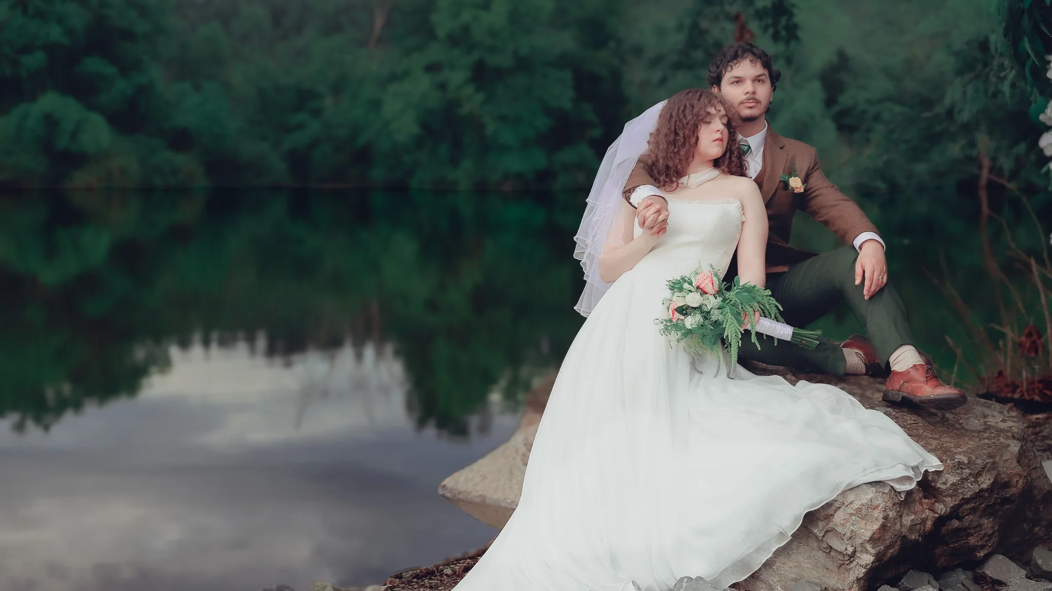 A newlywed couple sitting on a large rock beside a calm lake, surrounded by lush green trees, with the bride in a white wedding dress holding a bouquet, and the groom in a brown suit with dark pants, holding her hand.