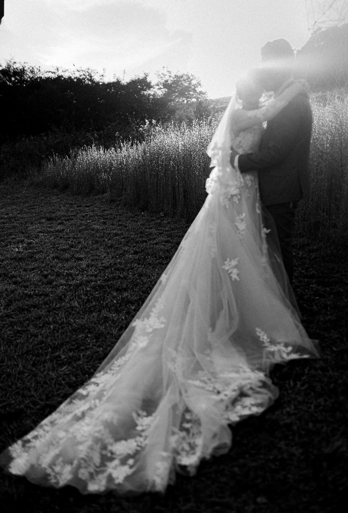 A bride and groom embracing outdoors at sunset or sunrise, with the bride's long lace train flowing behind them, on a grassy field with trees in the background.