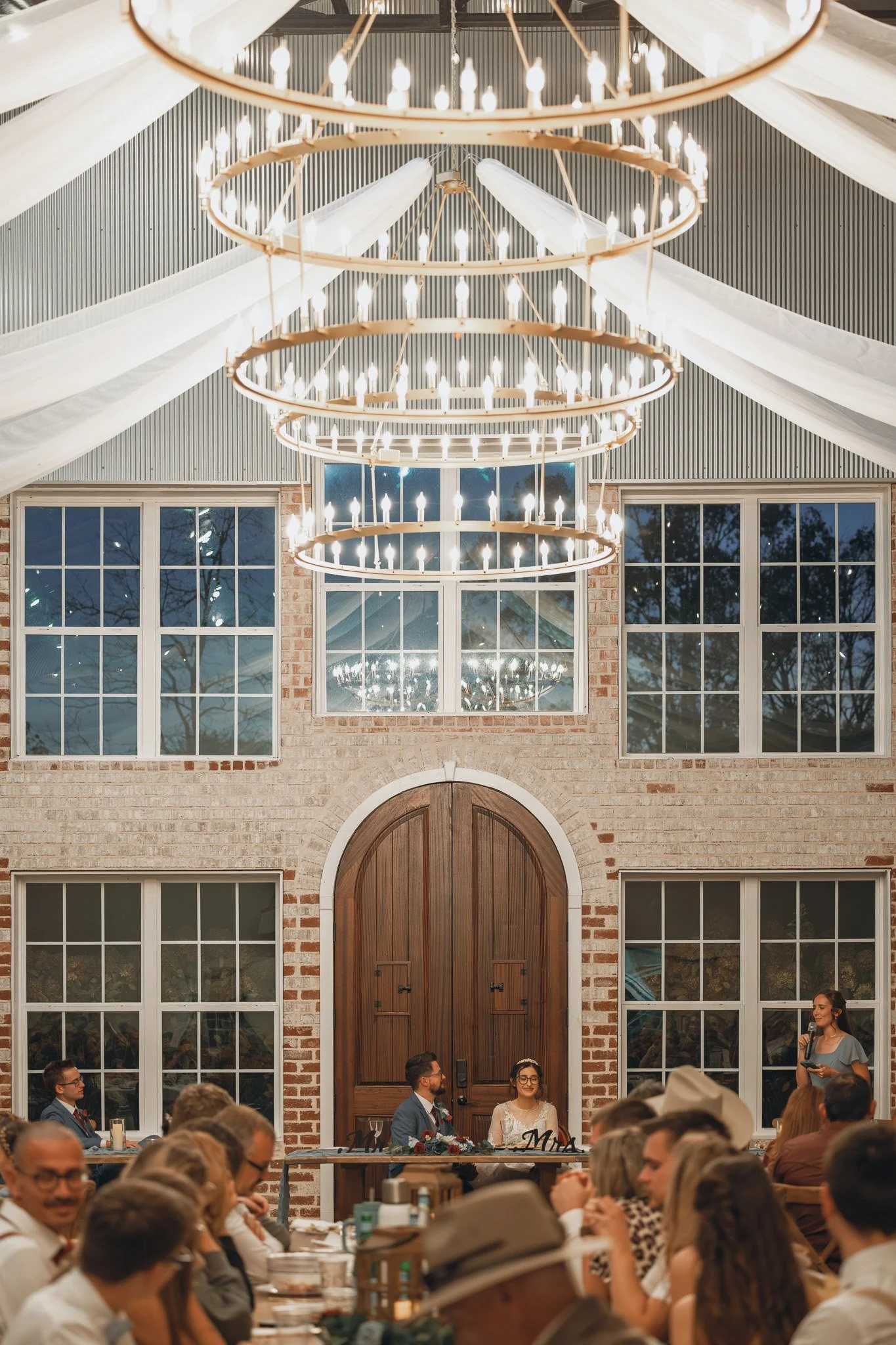 Indoor wedding reception with a brick wall, large wooden door, and big windows. Guests seated at tables, bride and groom sitting at a decorated head table, and a woman giving a speech. Large modern chandelier hanging from the ceiling with fabric drap