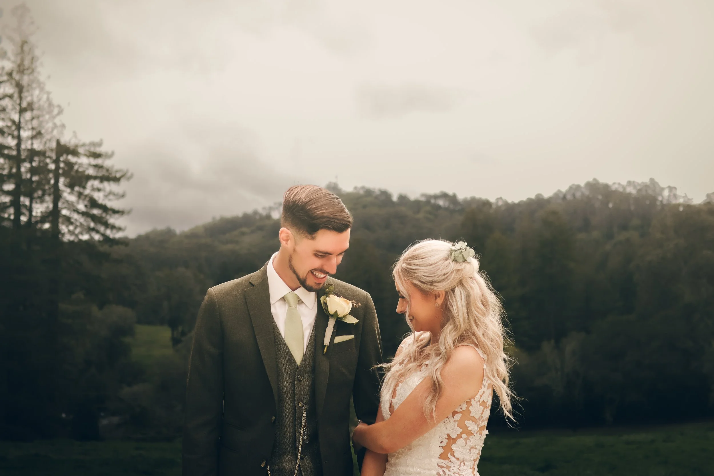 A bride and groom standing outdoors with trees and hills in the background, smiling and looking at each other.