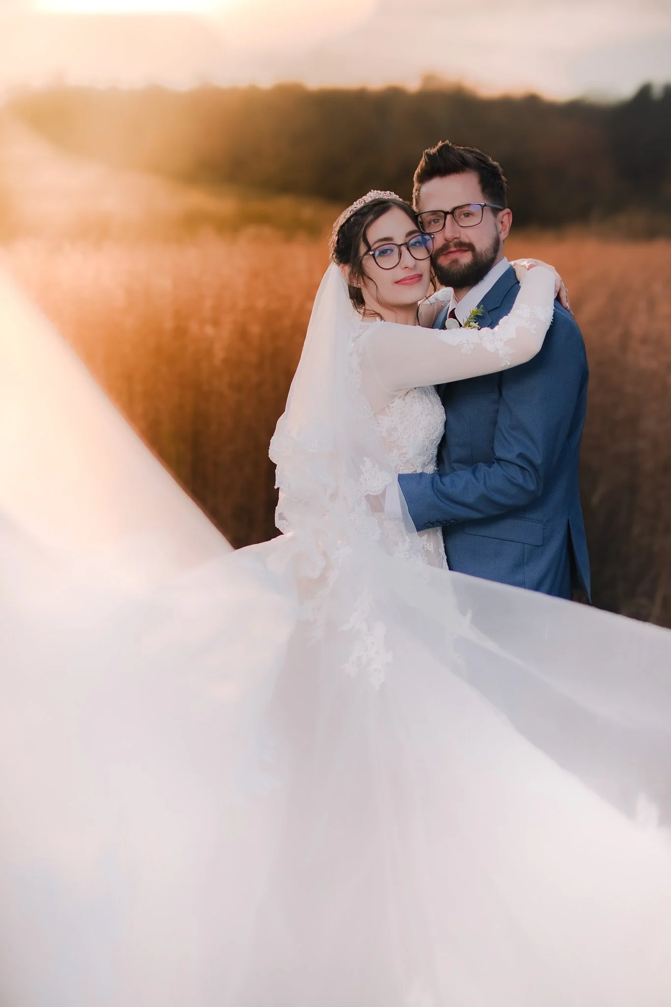 A bride and groom embrace outdoors during sunset, with a scenic natural background and the bride's flowing white wedding dress in the foreground.