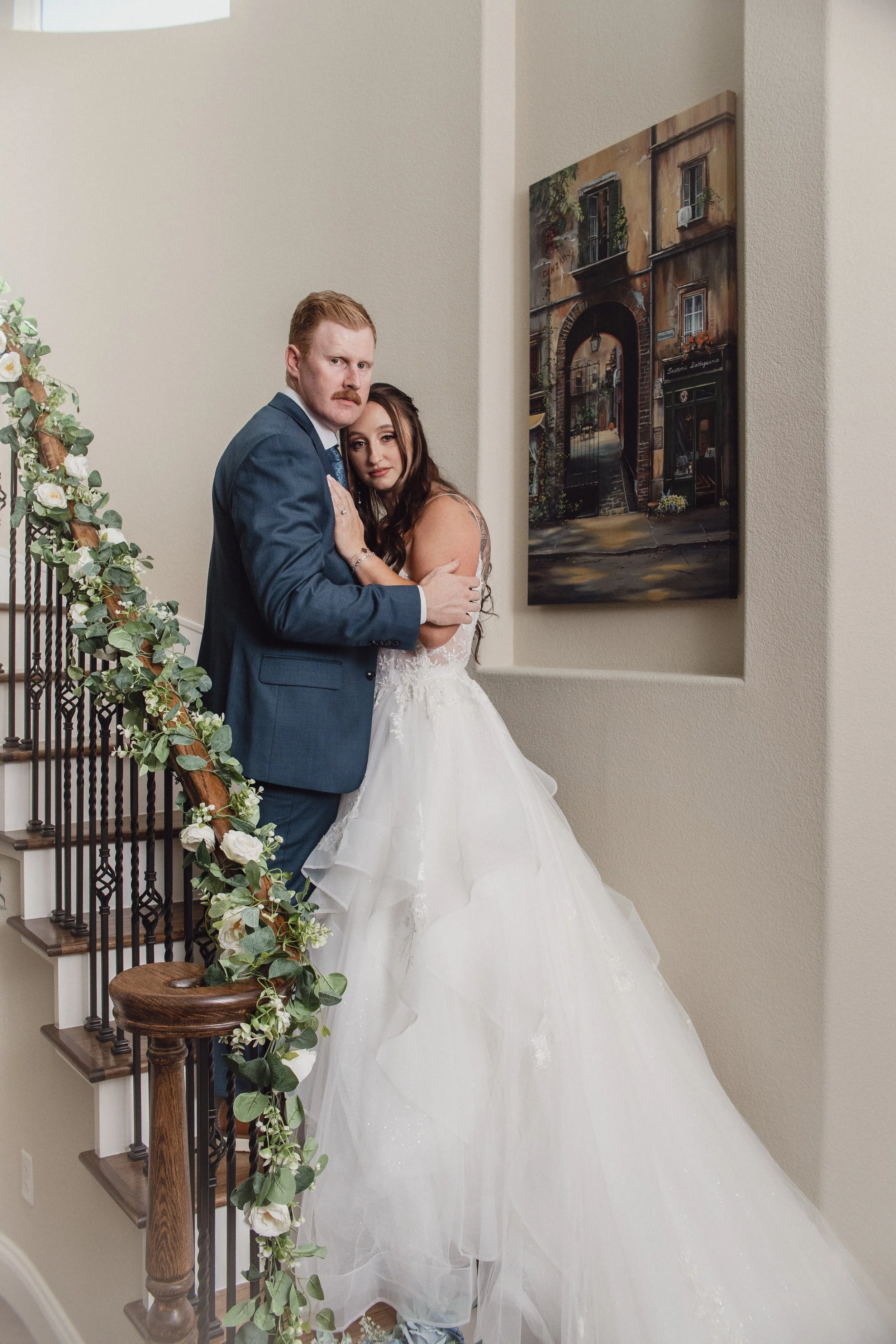 A couple dressed in wedding attire standing on staircase inside a house, with a painting of an alleyway on the wall.