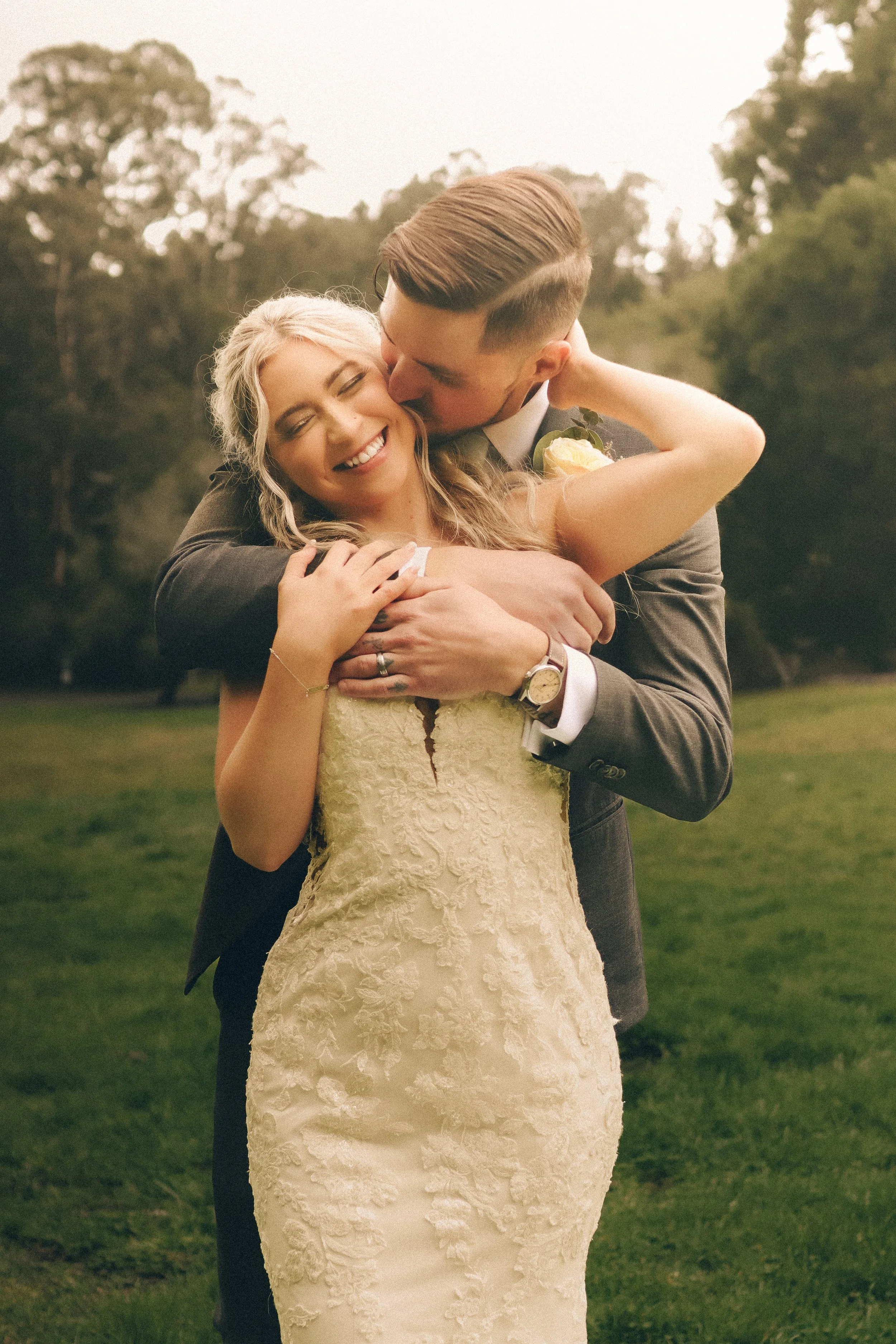 A smiling bride and groom embrace outdoors in a park, with the groom kissing the bride's forehead. The bride wears an elegant lace wedding gown, and the groom is dressed in a suit with a watch and a boutonniere.