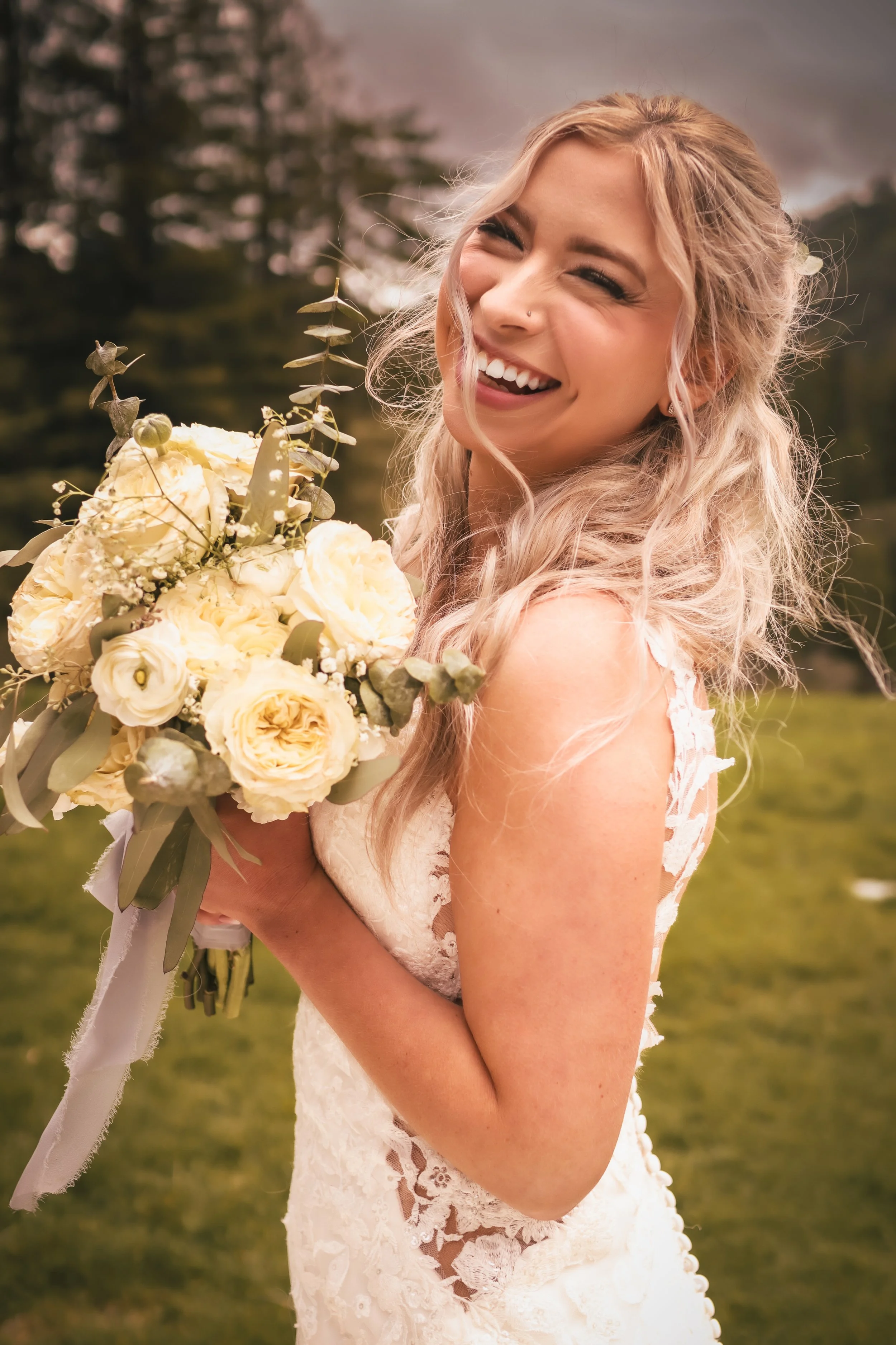A bride in a lace wedding dress holding a bouquet of white flowers, smiling outdoors with trees in the background.
