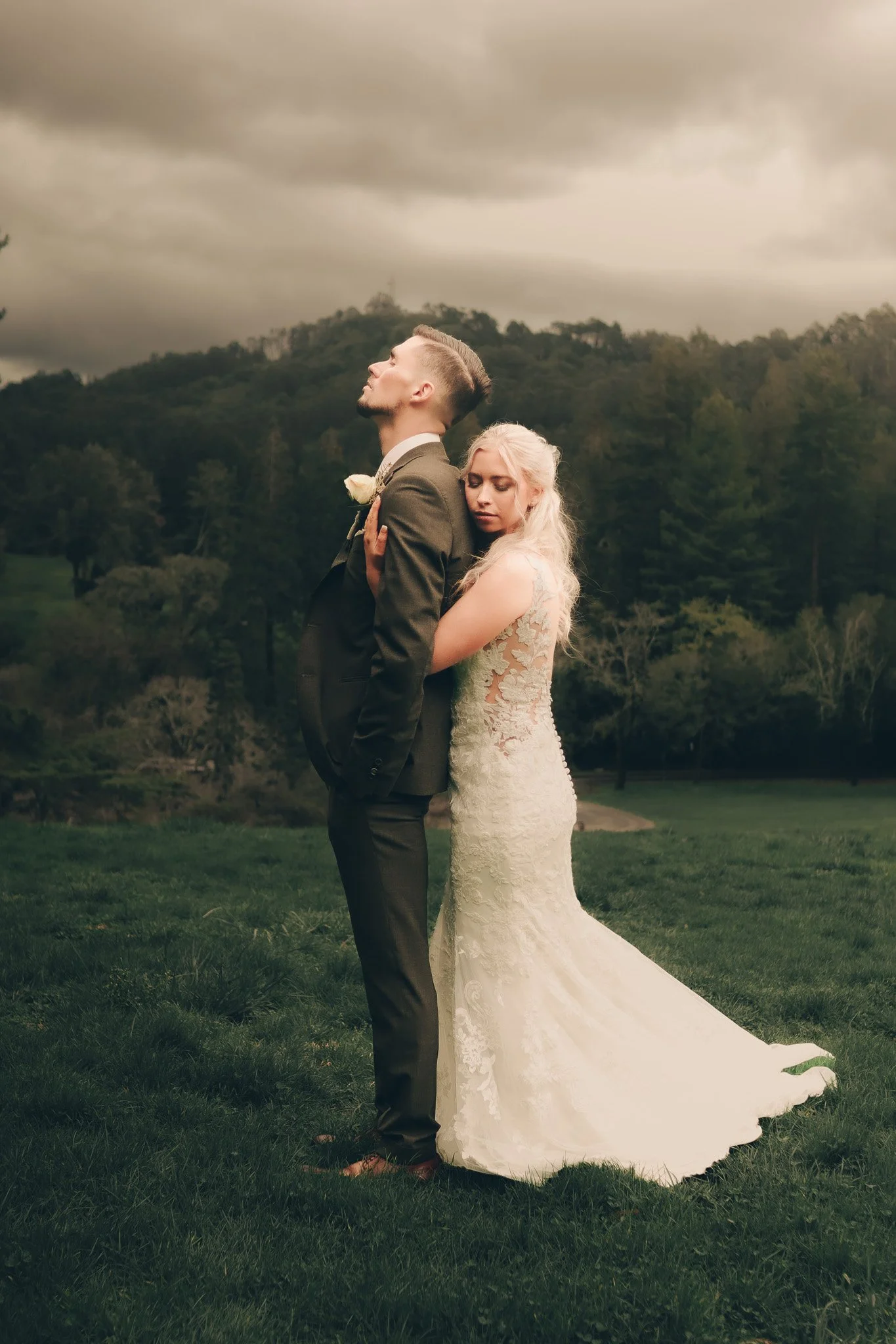 A bride and groom standing on grass outdoors, with the groom touching his chest and the bride resting her head on his shoulder, under a cloudy sky.