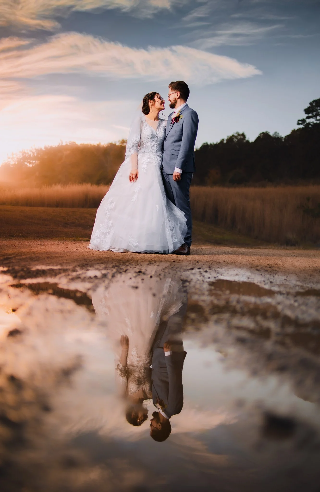 A bride and groom standing close together outdoors during sunset, with their reflection visible in a puddle on the ground.