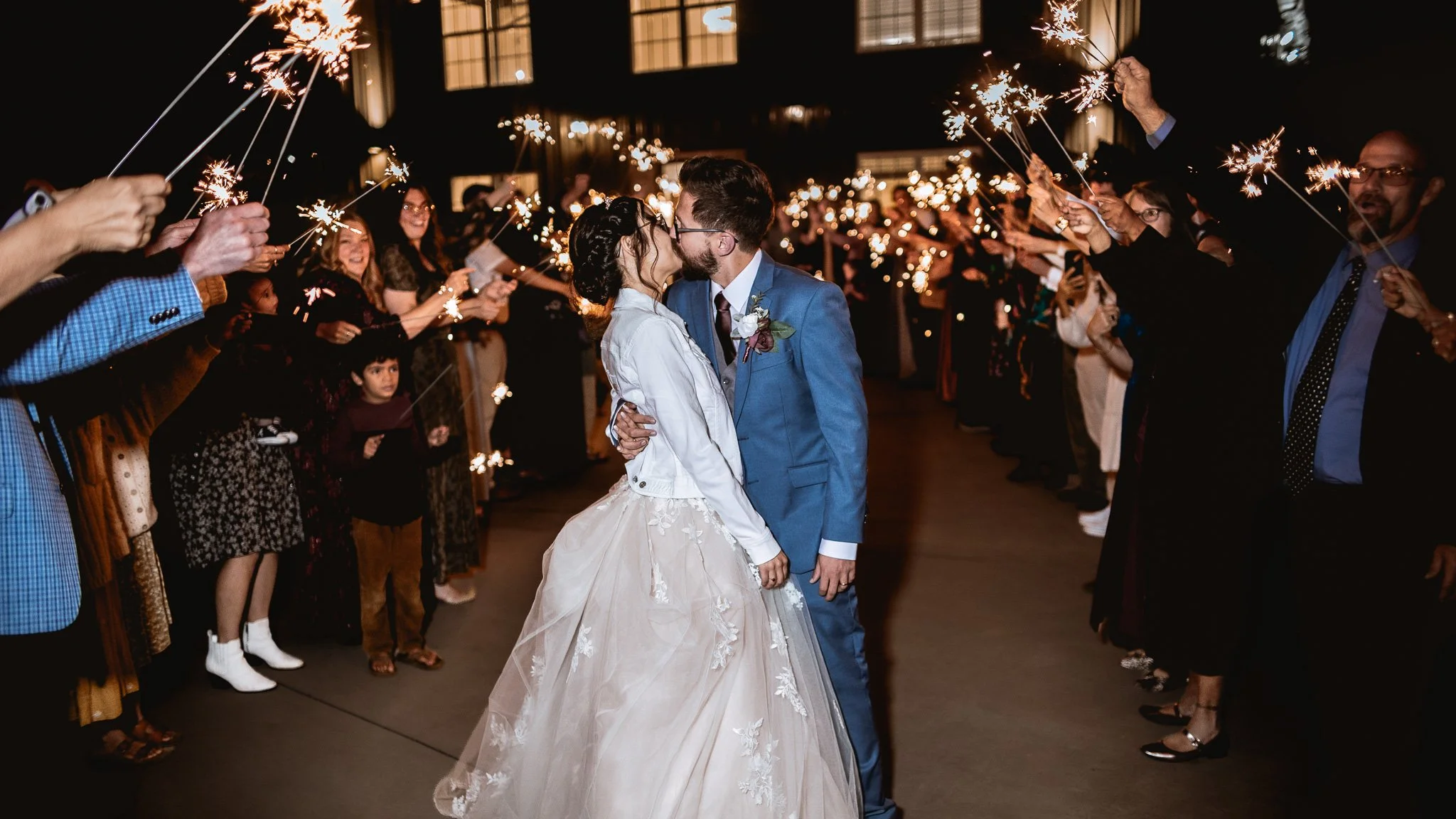 Couple sharing a kiss at their wedding reception surrounded by guests holding sparklers at night.
