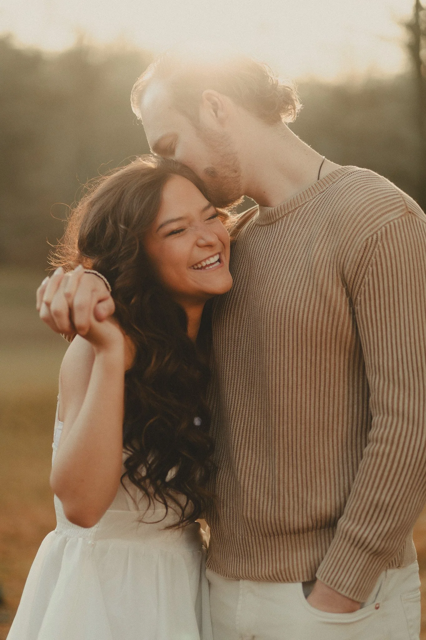 A happy couple embracing outdoors during golden hour, with the woman smiling and the man leaning his forehead against her head.