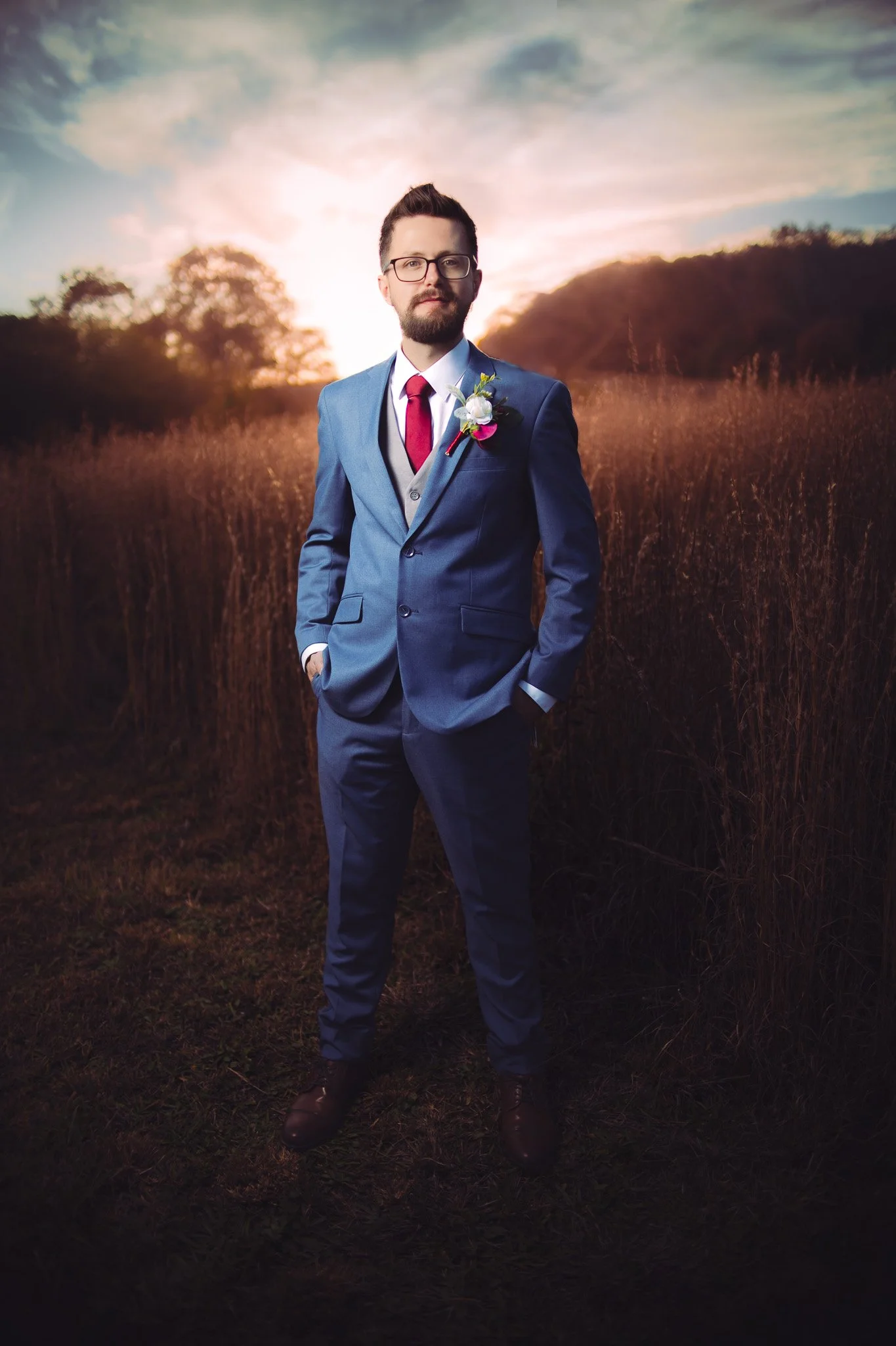 A man dressed in a blue suit with a red tie and boutonniere standing outdoors at sunset in a field of tall grass.