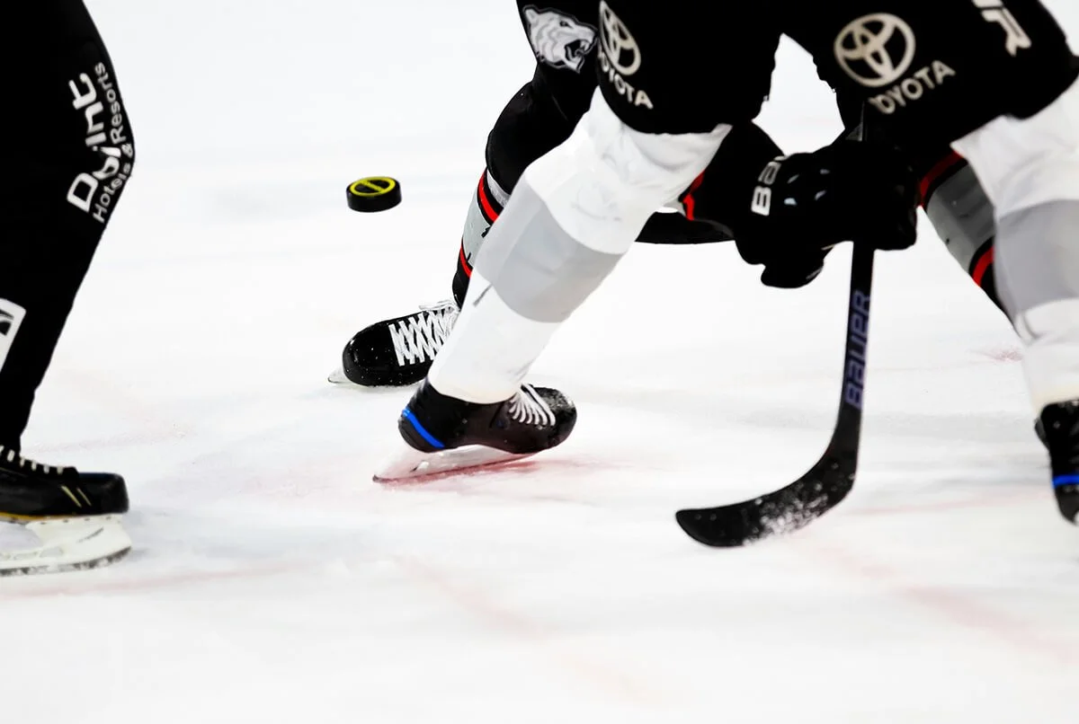 Two hockey players battling for puck on ice rink, one with hockey stick, puck mid-air, close-up shot.
