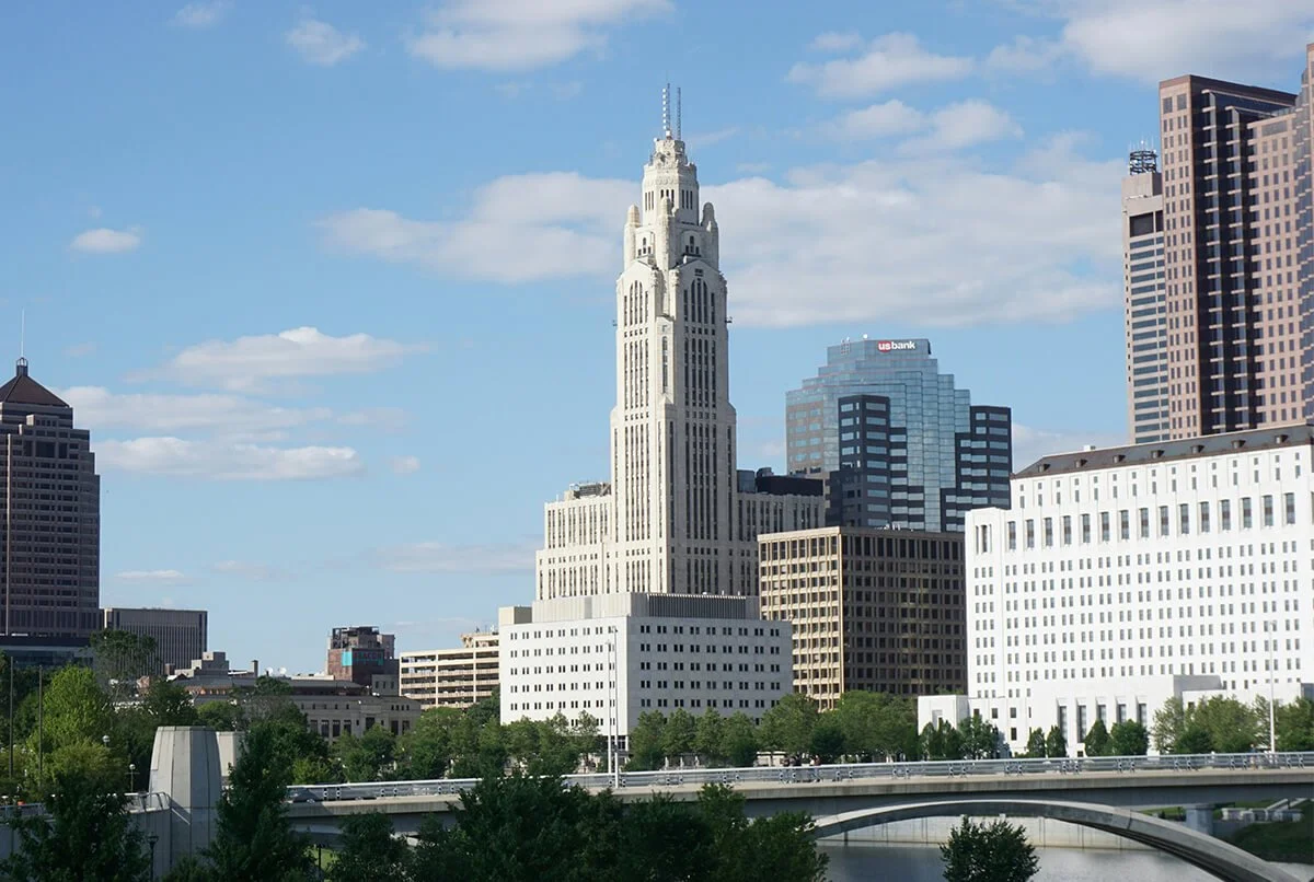 Columbus, Ohio city skyline featuring the tall, white LeVeque Tower, surrounded by various modern skyscrapers, with a bridge over a river in the foreground and partly cloudy blue sky.