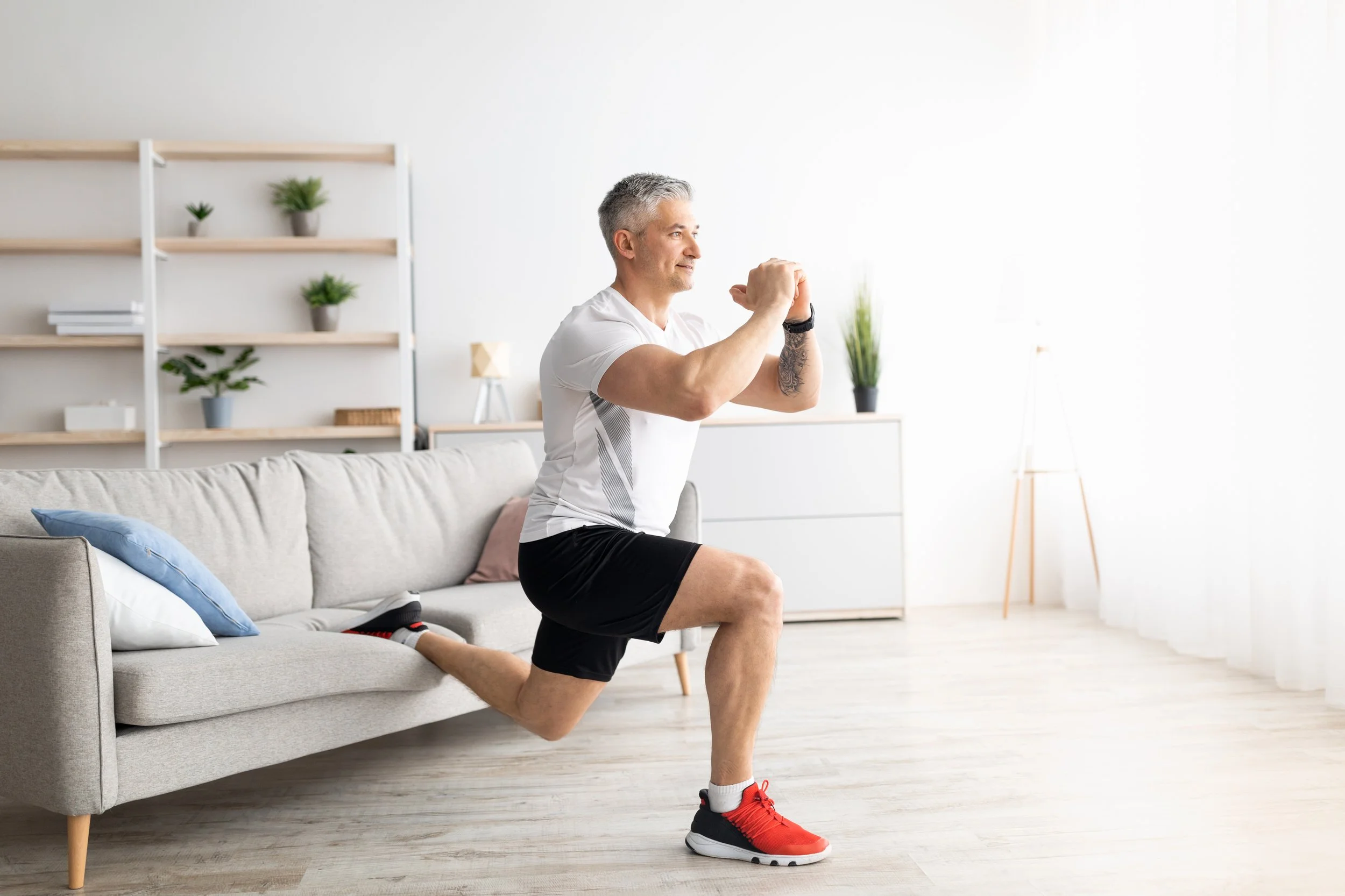 Man doing a lunge exercise in a bright, modern living room with a sofa and shelves with plants.