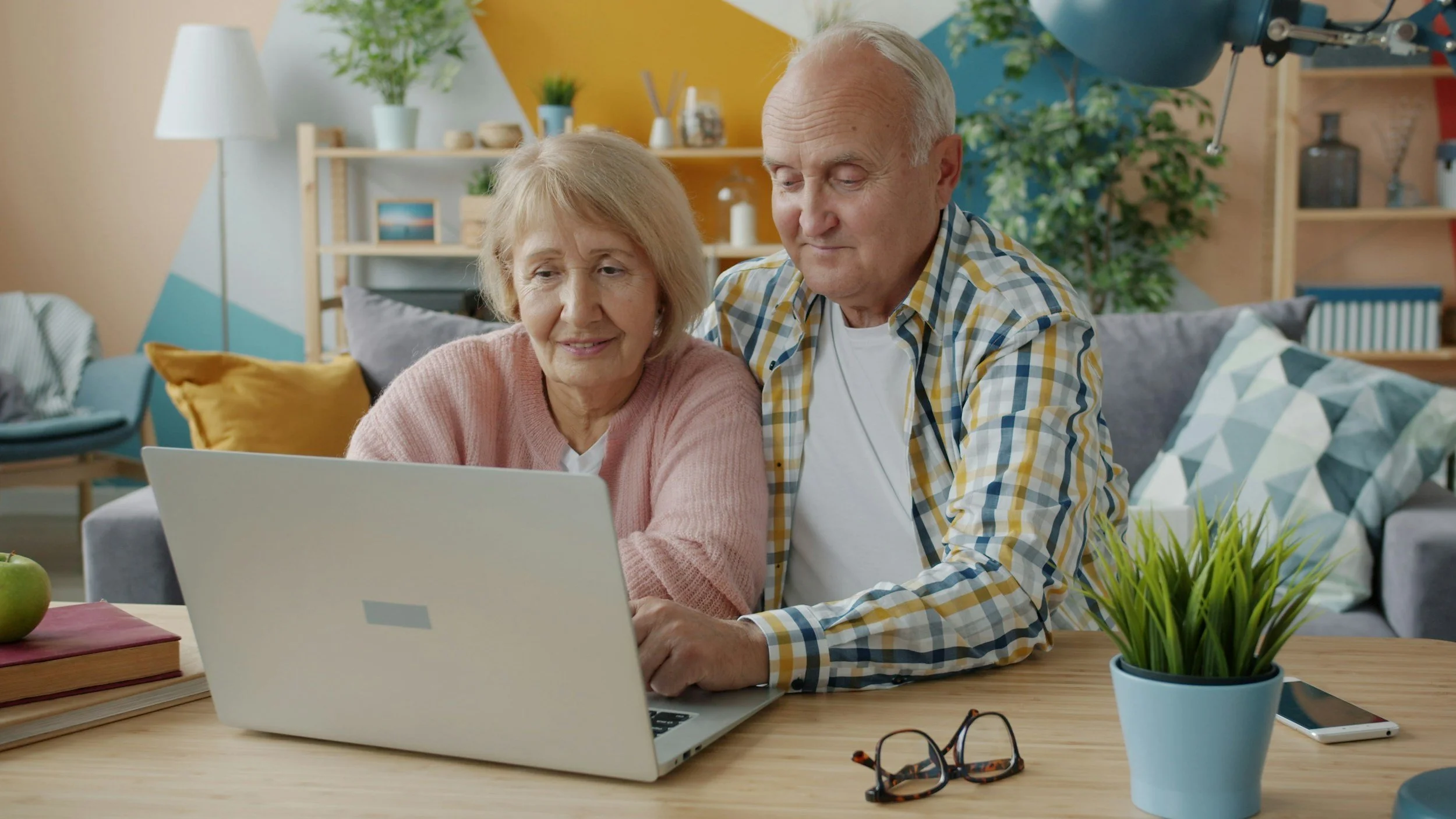 An elderly couple sitting at a wooden table, looking at a laptop together in a brightly decorated living room with plants, books, and colorful pillows.