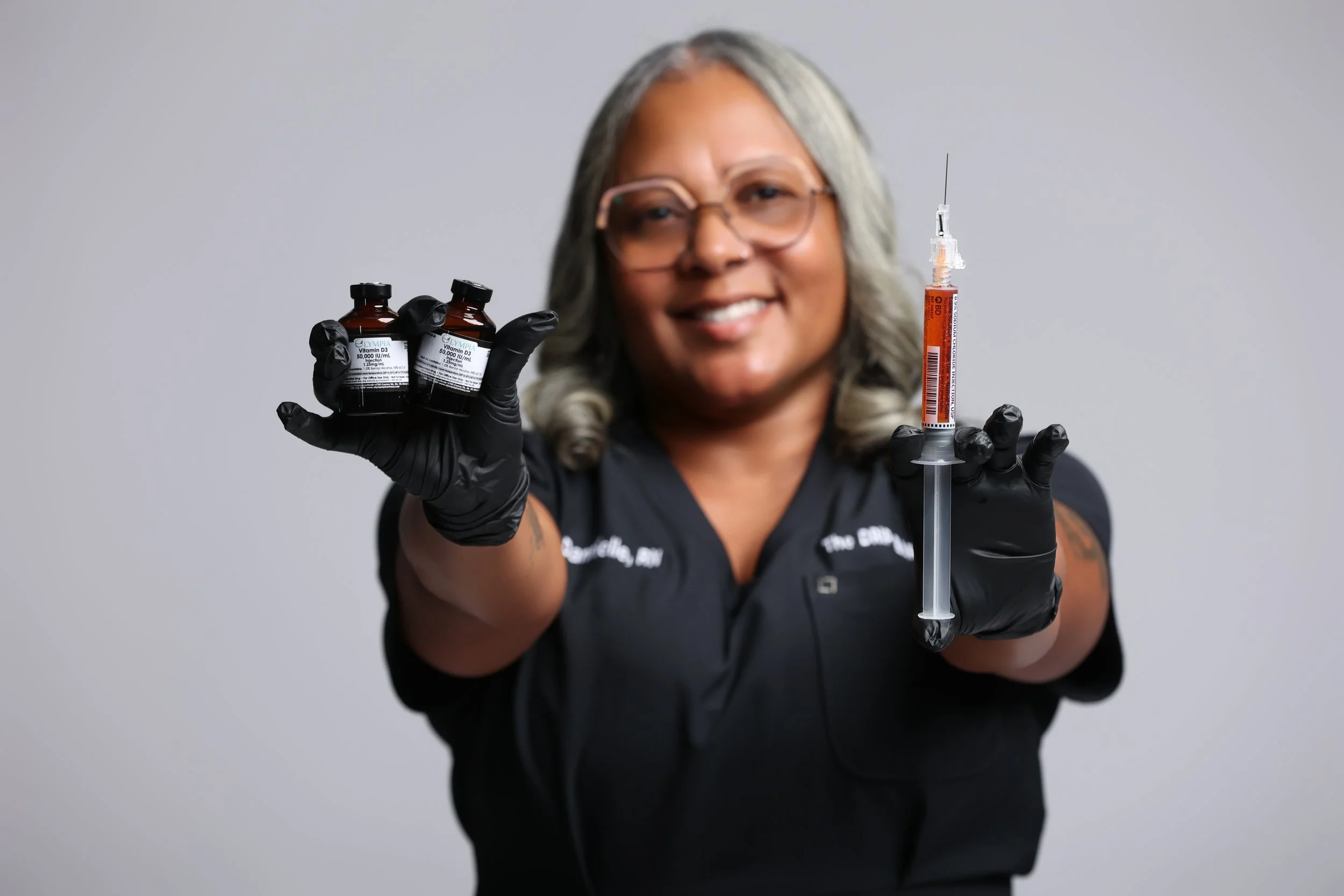 A smiling woman wearing glasses and black gloves holding two vials of medicine in one hand and a syringe in the other, against a plain gray background.