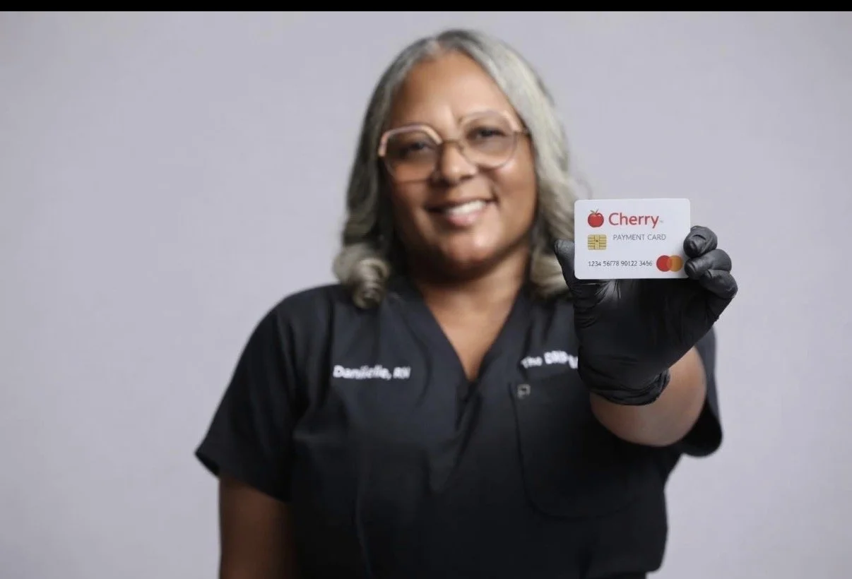 A woman with gray hair and glasses smiling, wearing a black uniform, holding a white credit card with a cherry logo near her face.