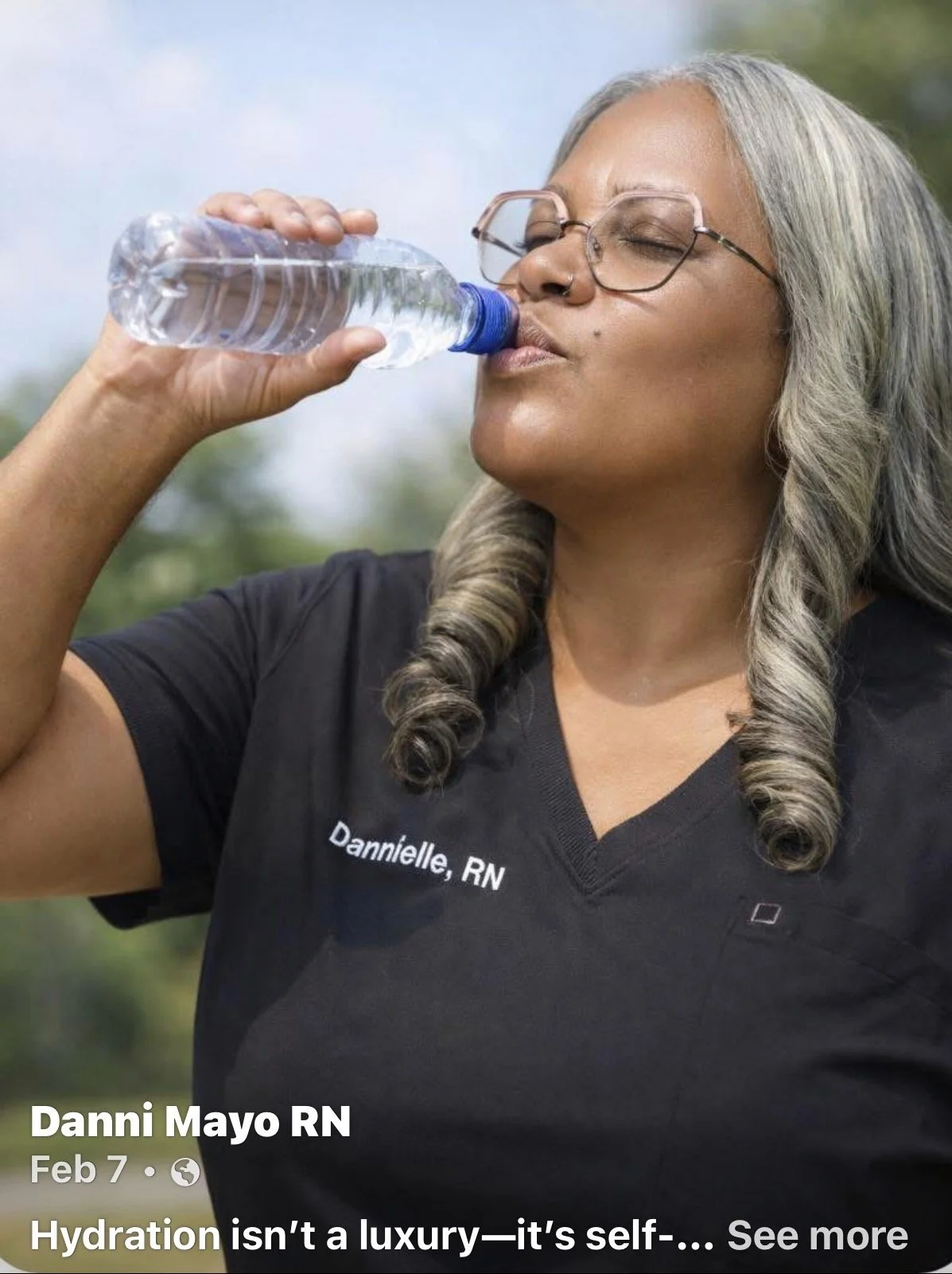 A woman with gray hair, glasses, and a nose ring drinking water from a plastic bottle outdoors. She is wearing a black shirt with the name tag "Danni Mayo RN".