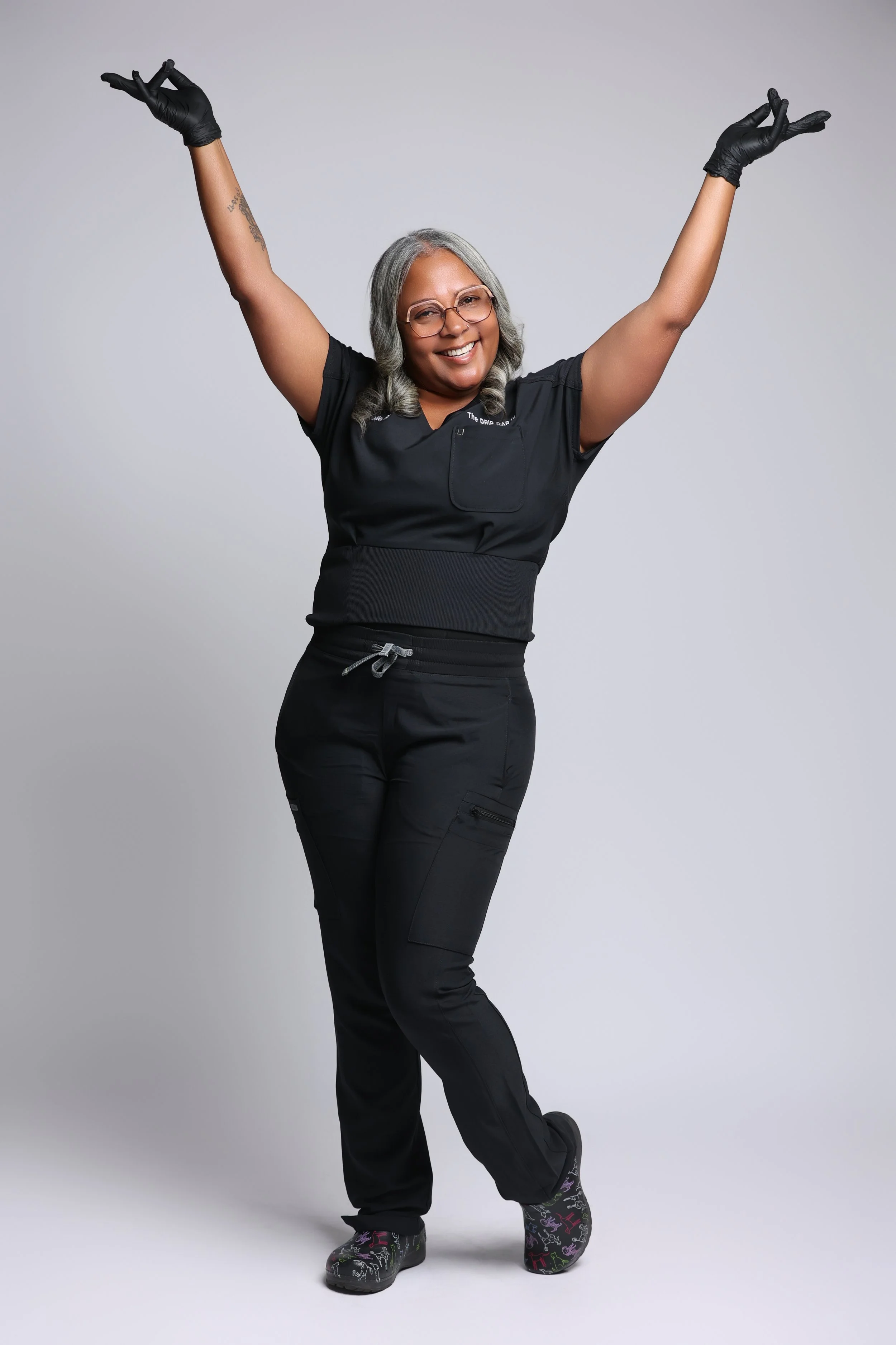 A woman with gray hair, glasses, and a big smile, wearing black medical scrubs and black gloves, standing with her arms raised in a joyful gesture, against a light gray background.