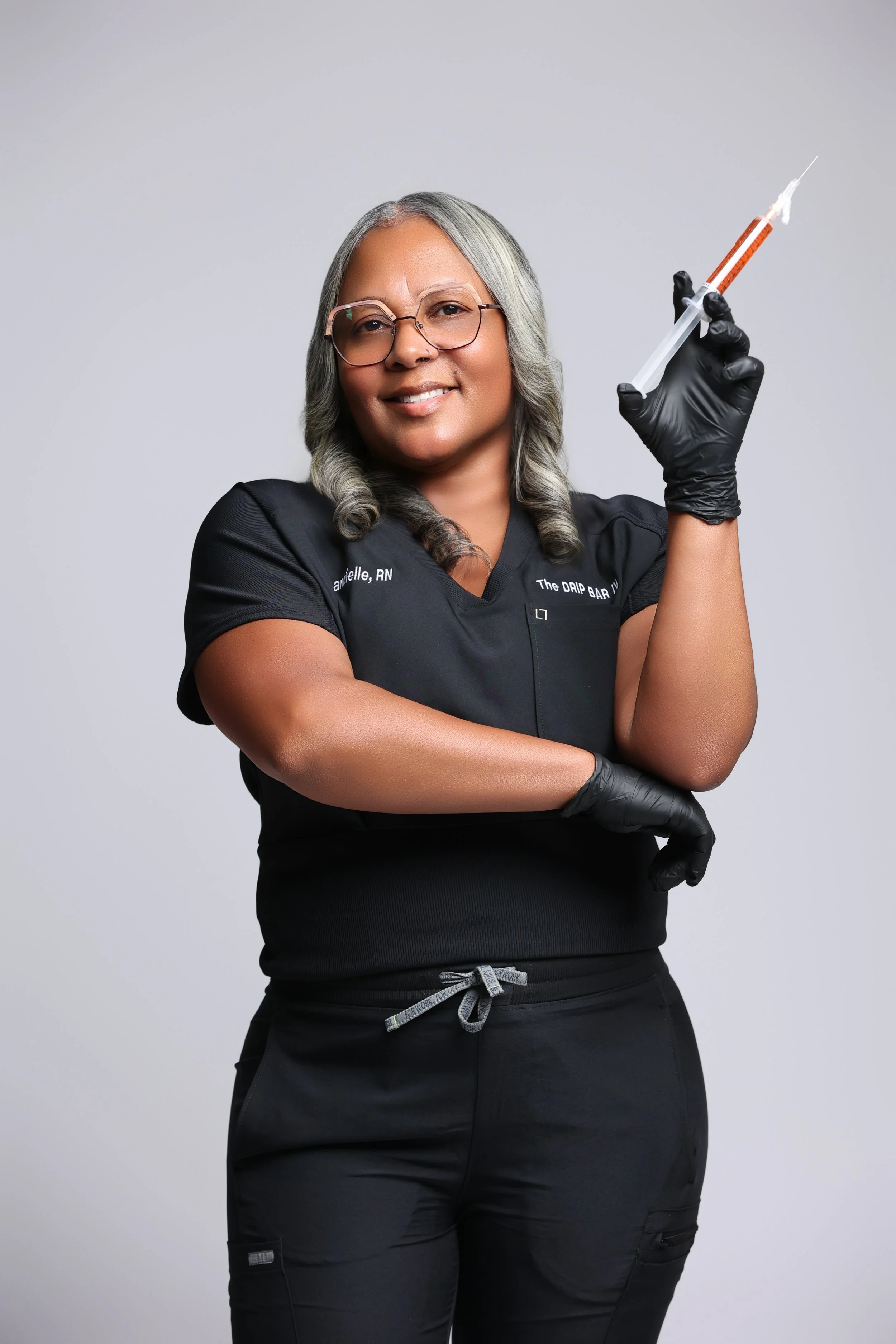 A smiling female nurse or medical professional with gray hair, glasses, and a nose ring, wearing black scrubs with her name and title, holding a syringe with orange liquid in her right hand, wearing black gloves, against a plain gray background.