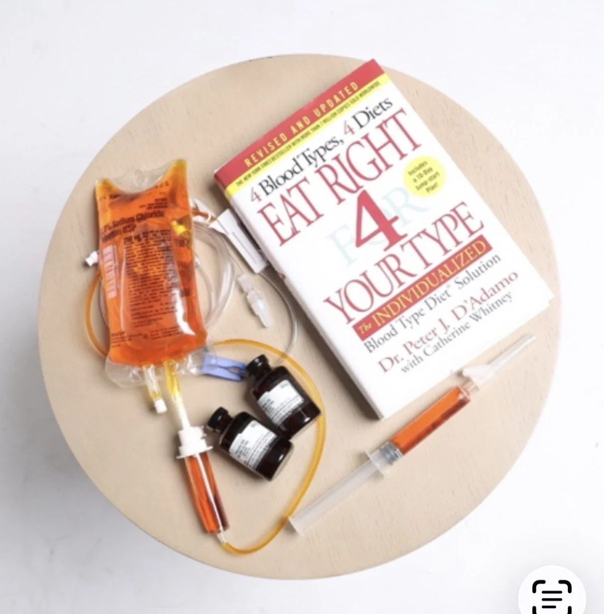 A blood donation setup with IV drip, blood bags, medication bottles, and a book titled 'Eat Right 4 Your Type' on a round wooden table.