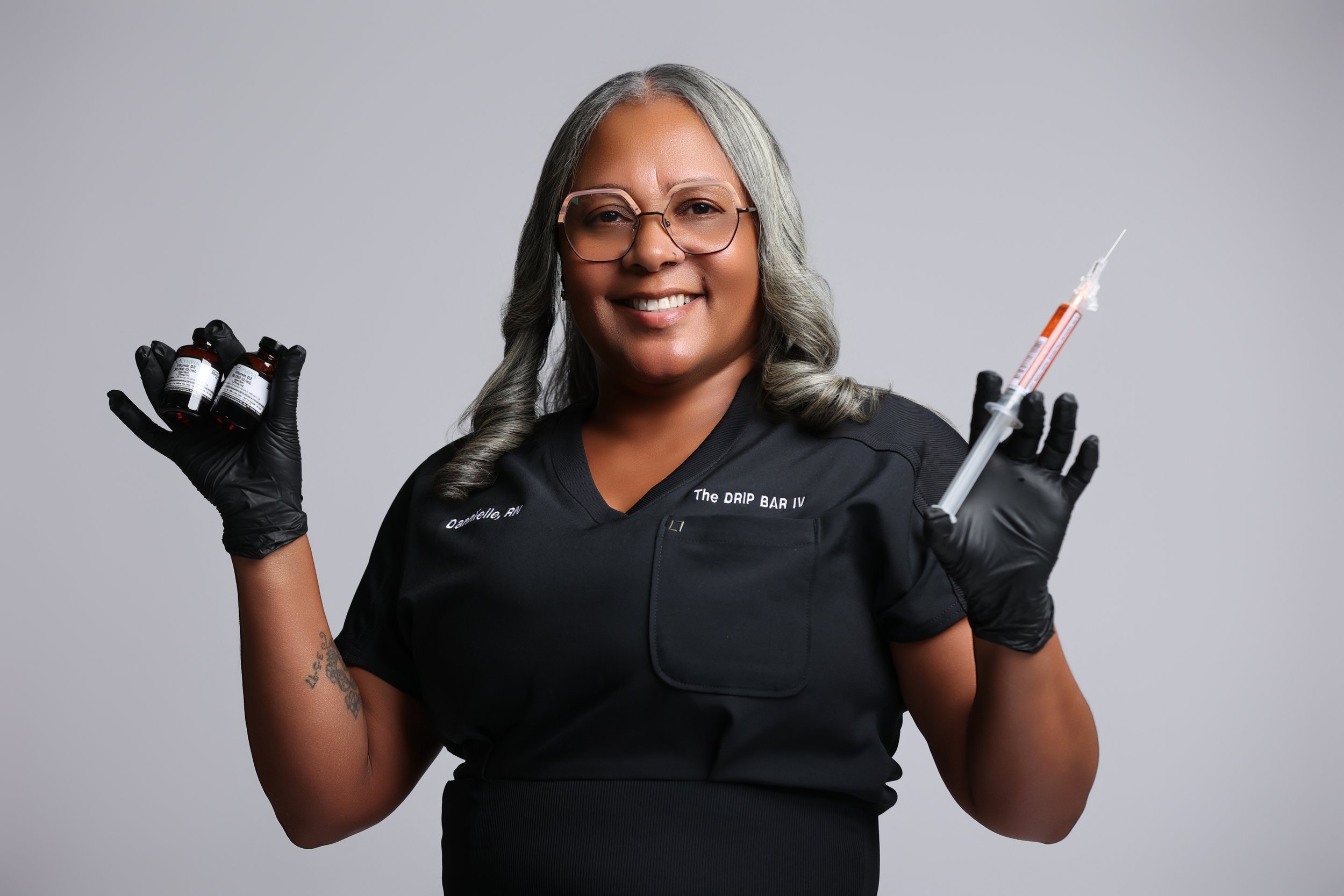 A smiling woman with gray hair and glasses holding bottles of medicine and a syringe, wearing black gloves and a black medical uniform, against a plain gray background.