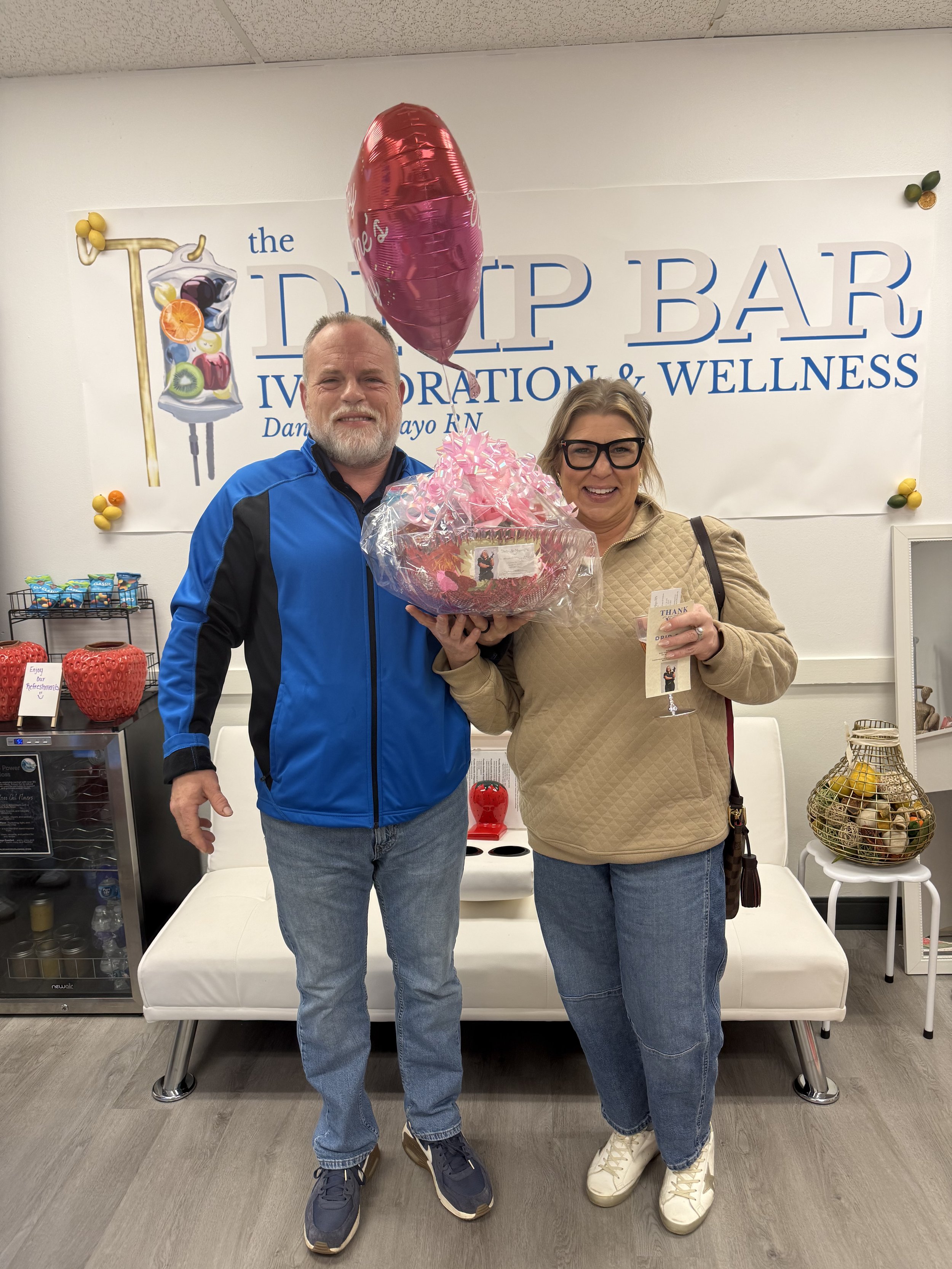 A man and a woman standing indoors in front of a sign that says 'the DIP BAR, IV Nutrition & Wellness.' They are smiling, and the woman is holding a gift basket wrapped in clear cellophane with pink ribbons and a pink balloon attached. The man is wearing a blue jacket and jeans, and the woman is wearing glasses, a beige jacket, and jeans.
