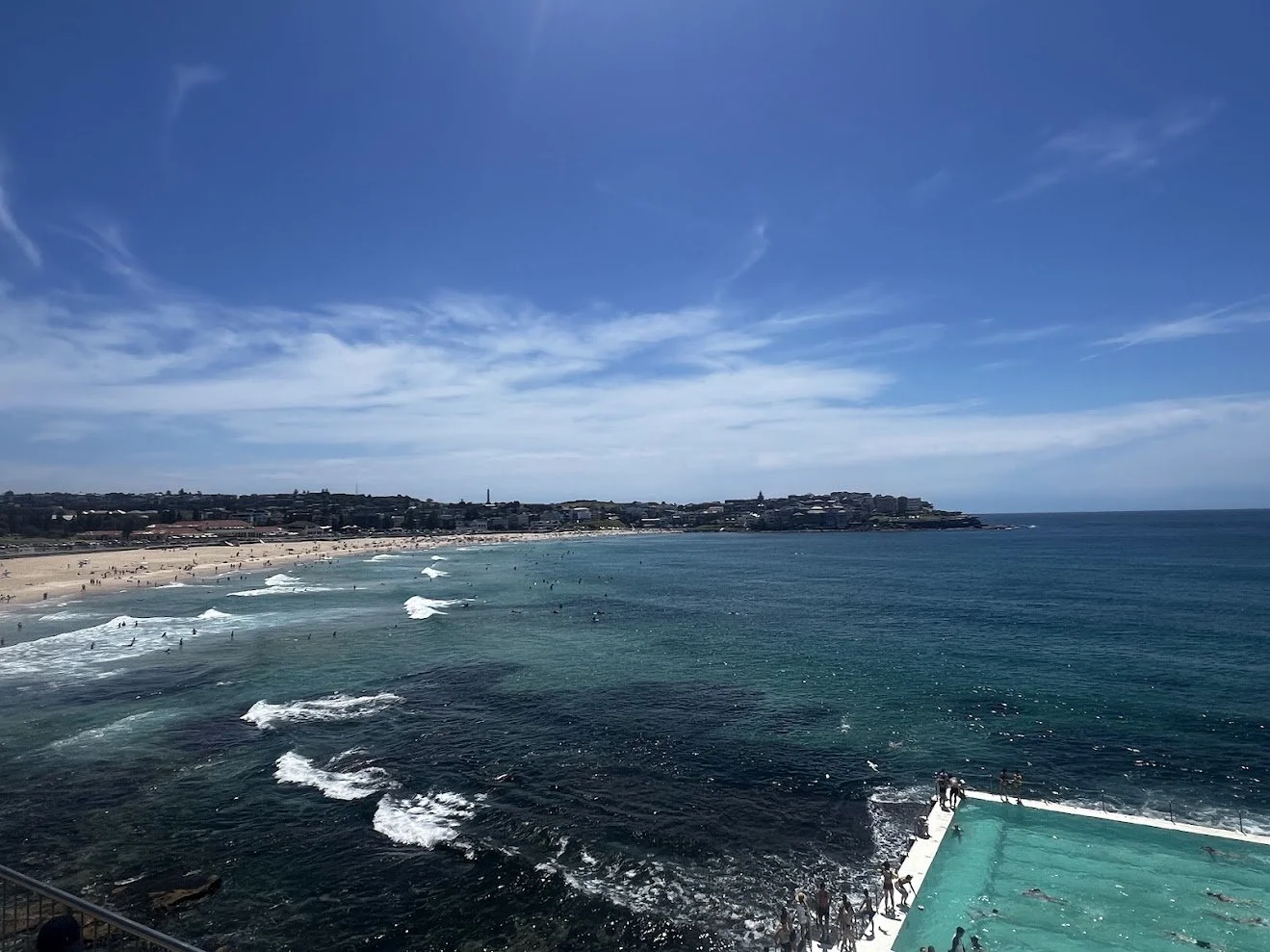 View of a crowded beach with people swimming and surfing, a swimming pool in the foreground, and a cityscape on a sunny day with a blue sky and scattered clouds.