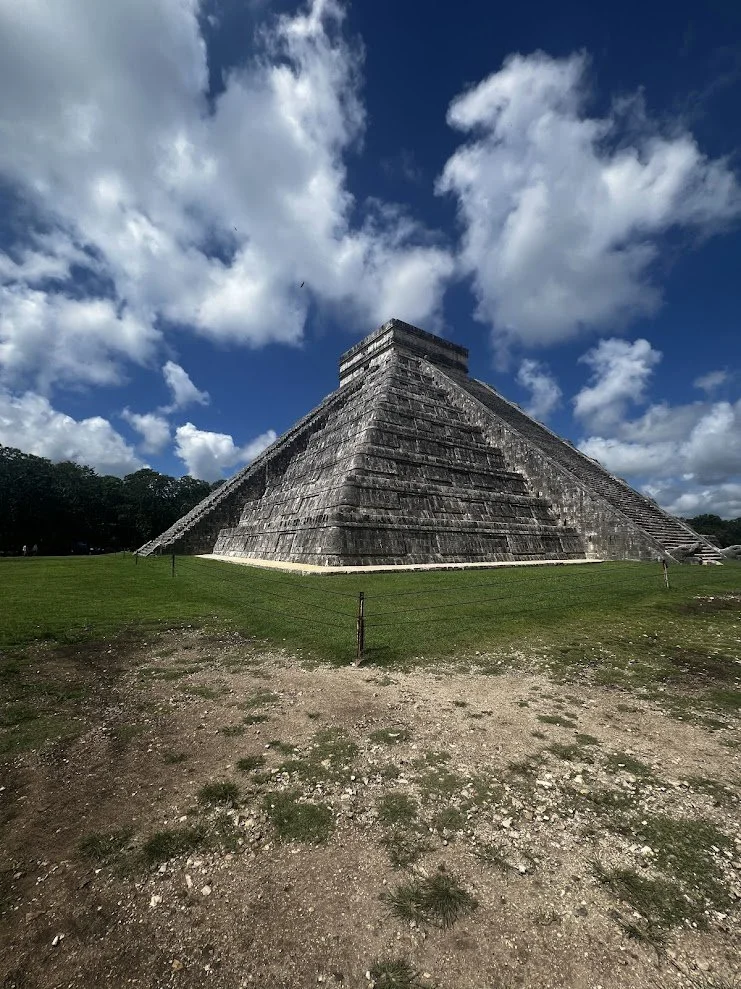 A large stepped pyramid, known as El Castillo at Chichen Itza, with a bright blue sky and white clouds in the background.
