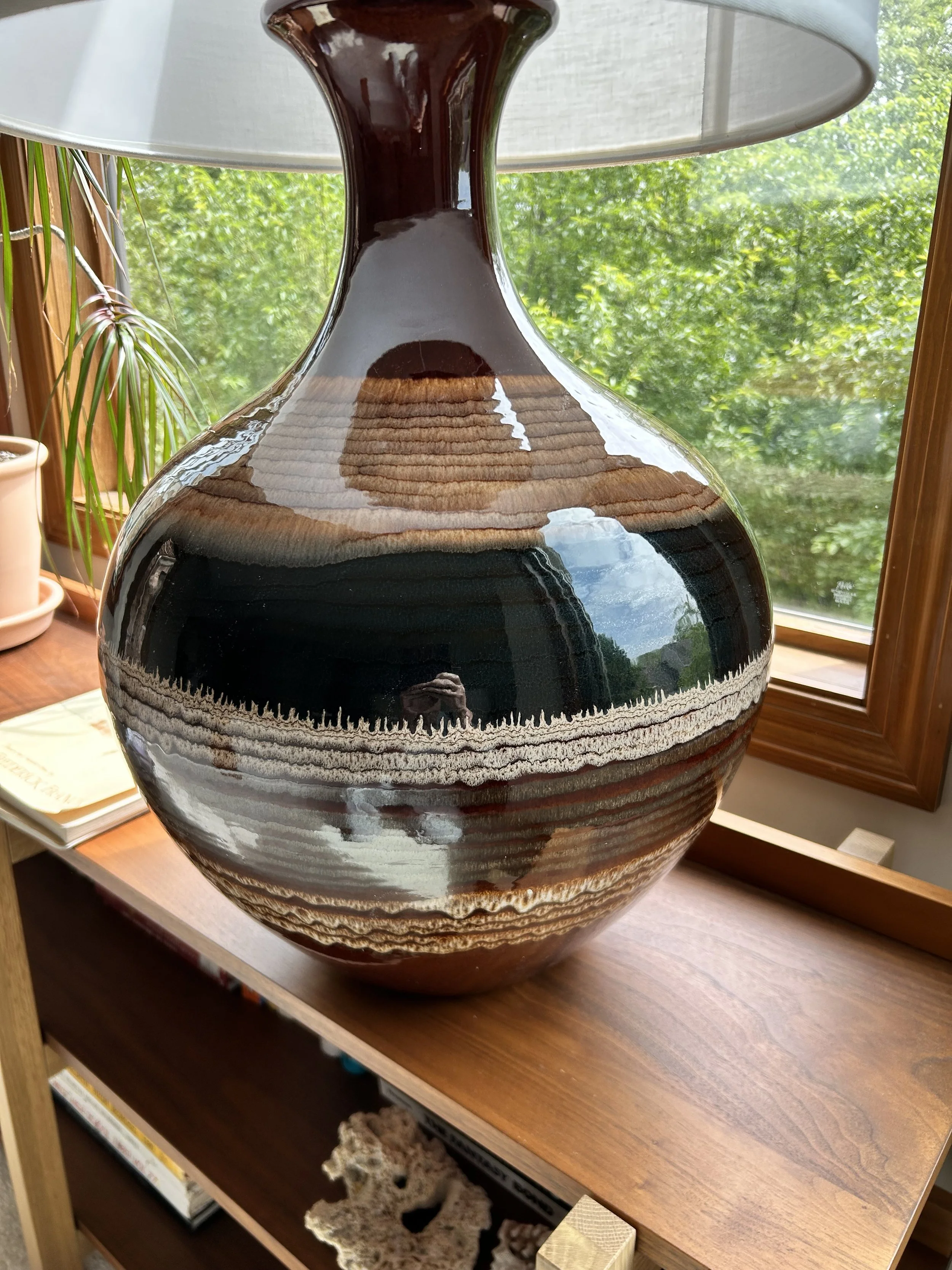 A large ceramic vase with a glossy, marbled pattern of brown, beige, and black sits on a wooden table near a window with green trees visible outside. A lamp with a white shade is positioned above the vase.