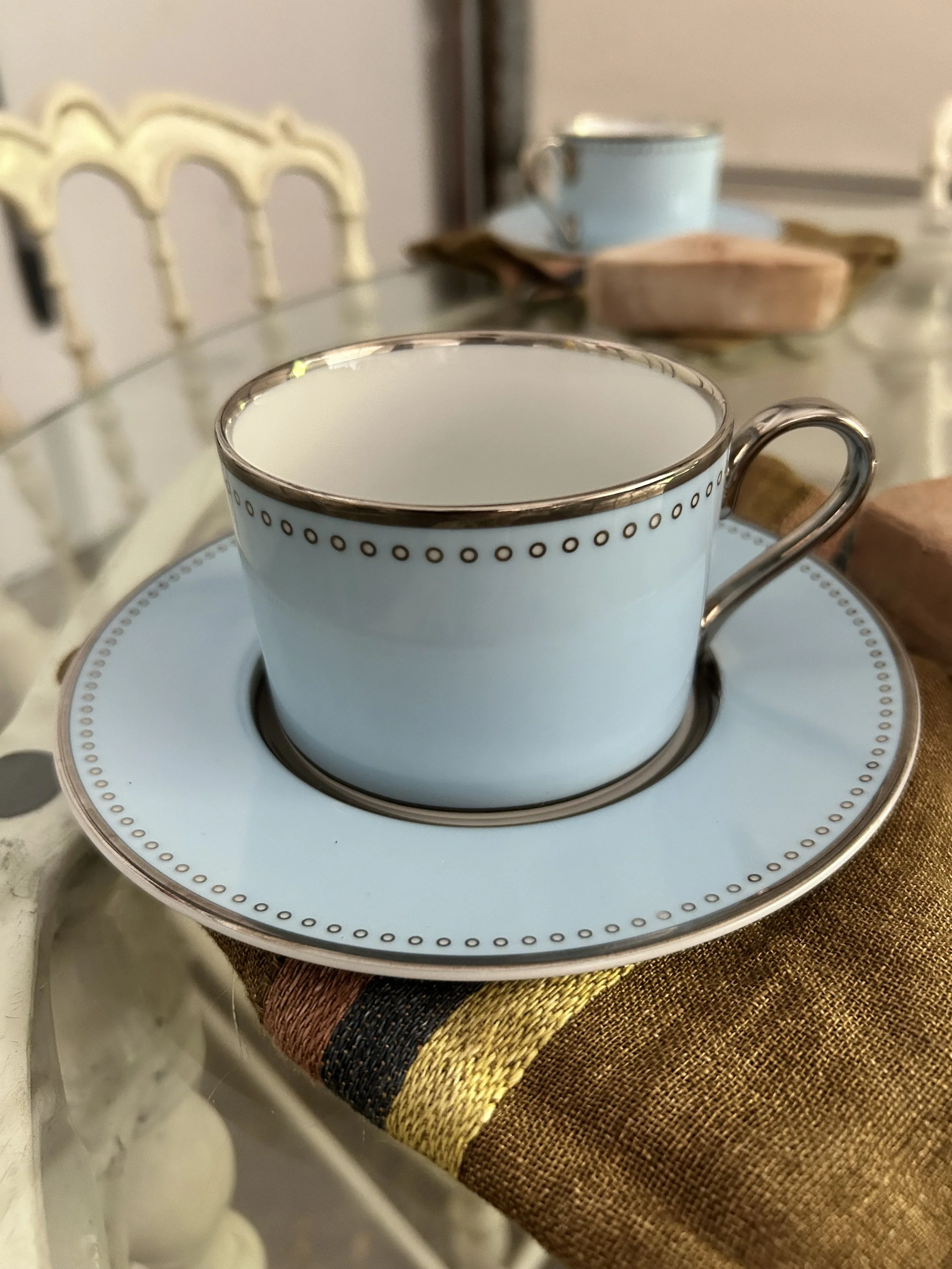 A light blue ceramic teacup with gold and black decorative trim on the rim, placed on a matching saucer, on a brown and yellow striped cloth. In the background, there is a second similar teacup and saucer on a table.