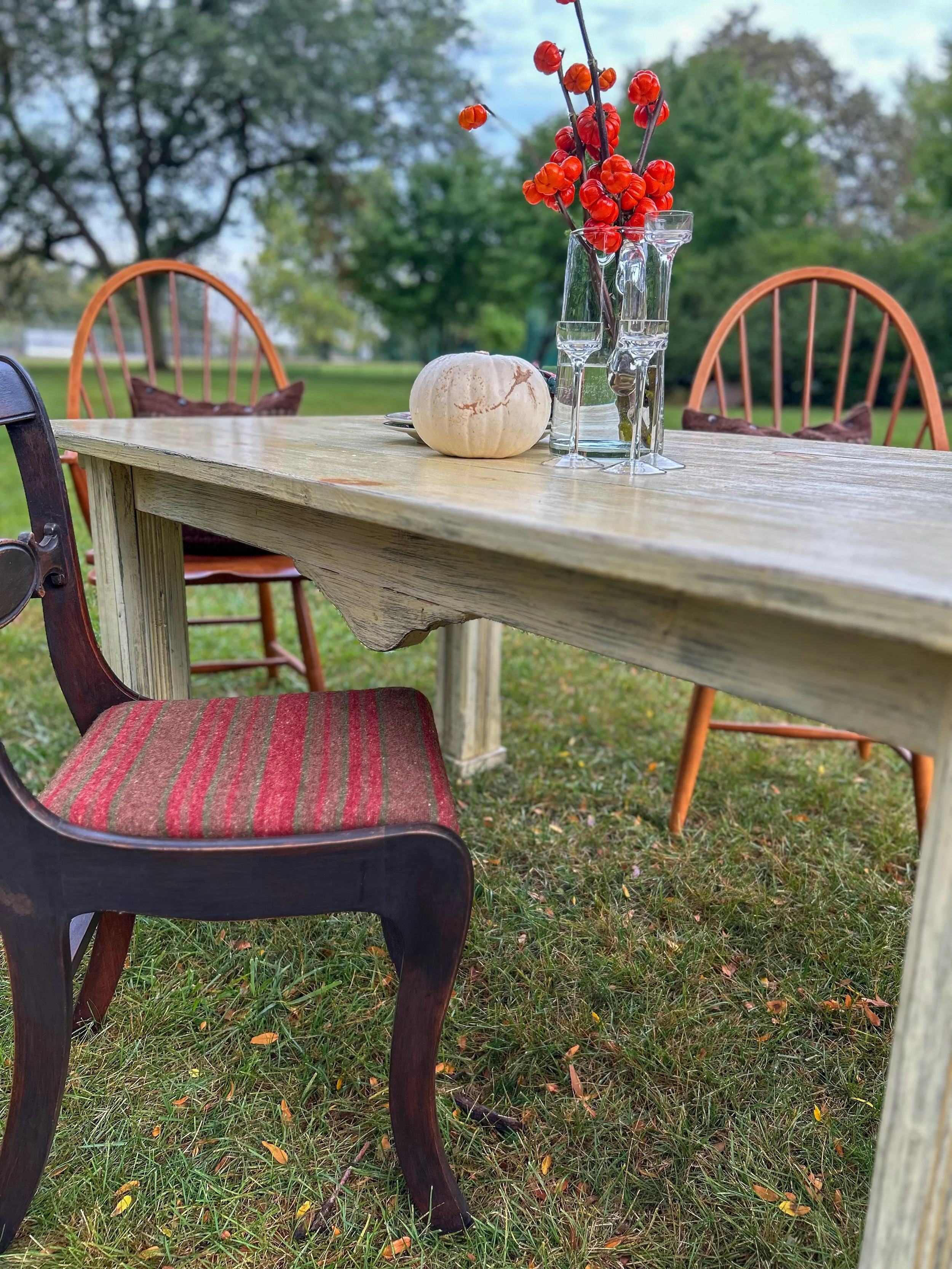 An outdoor dining setup with a wooden table, vintage chairs, and decorative items including a white pumpkin, glass vases, and a bouquet of red berries, set on a grassy lawn with trees in the background.