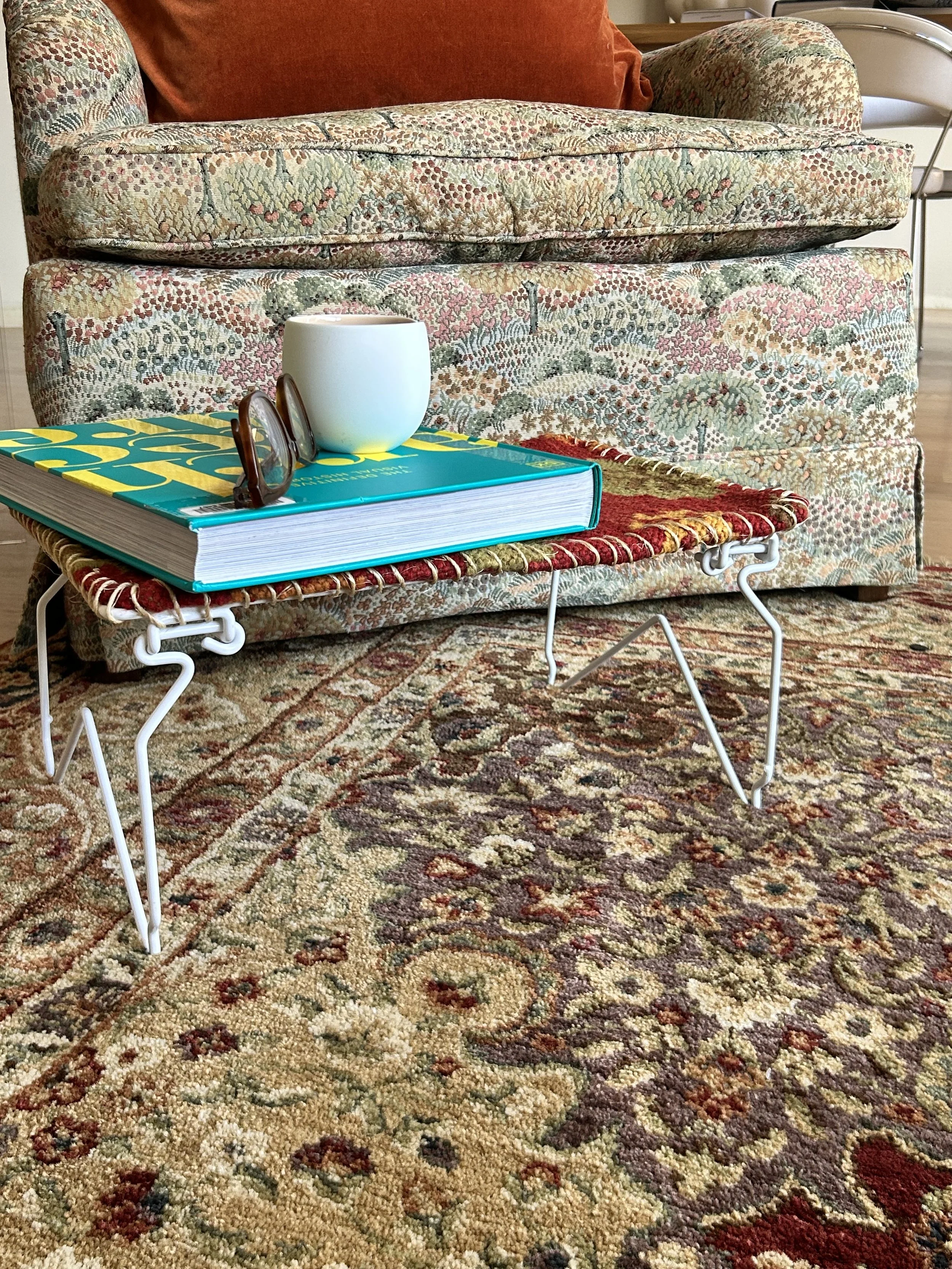 A cozy living room scene with a patterned fabric sofa, a small colorful woven table with a book, a white mug, and a pair of glasses on top, and a detailed patterned rug on the wooden floor.