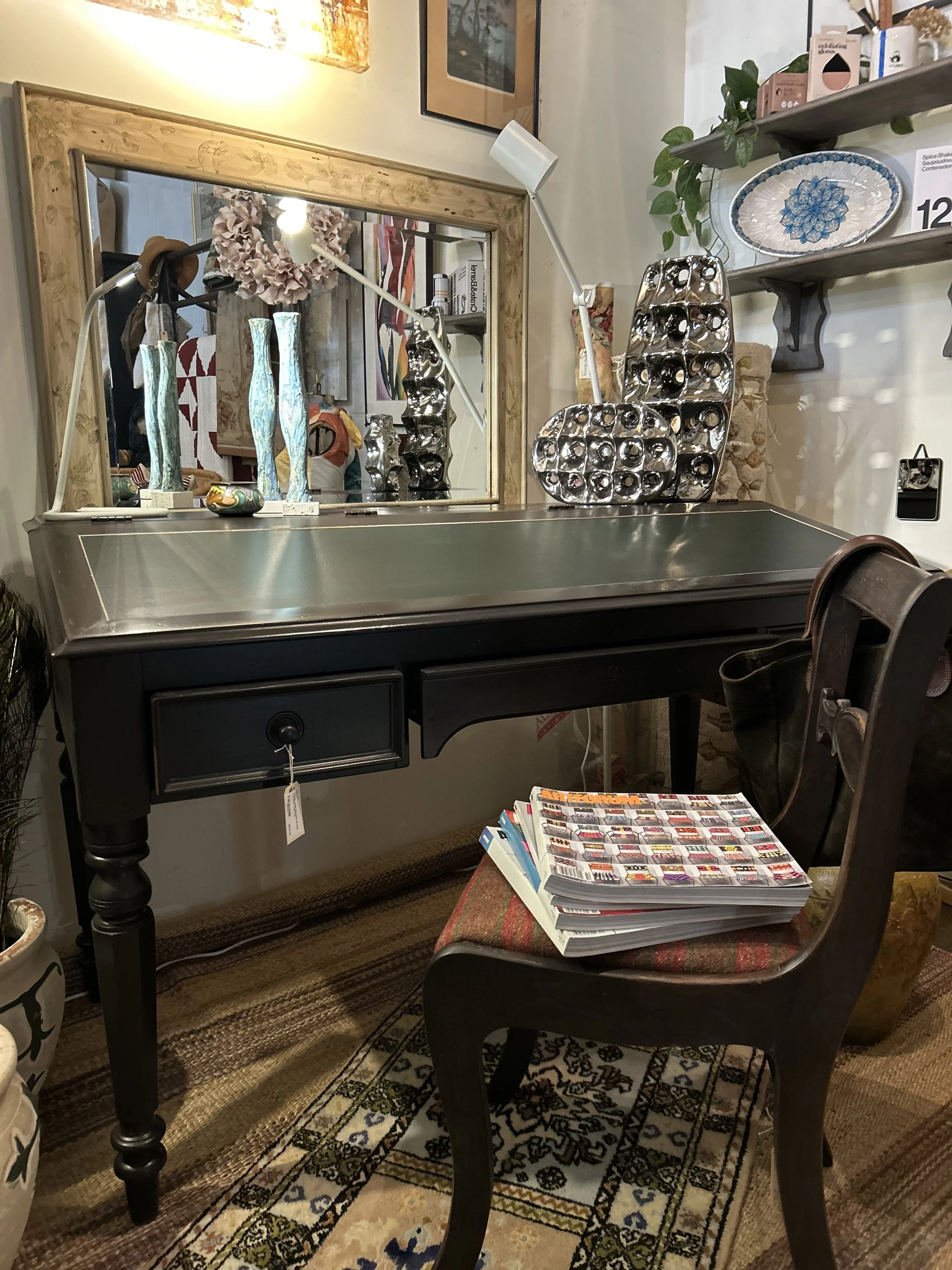 A vintage black writing desk with a metallic top, a drawer, and turned legs. On top are modern decorative silver vases and a lamp. Behind the desk is a mirror, reflecting some decorative items and artworks. To the right is a chair with a stack of mag
