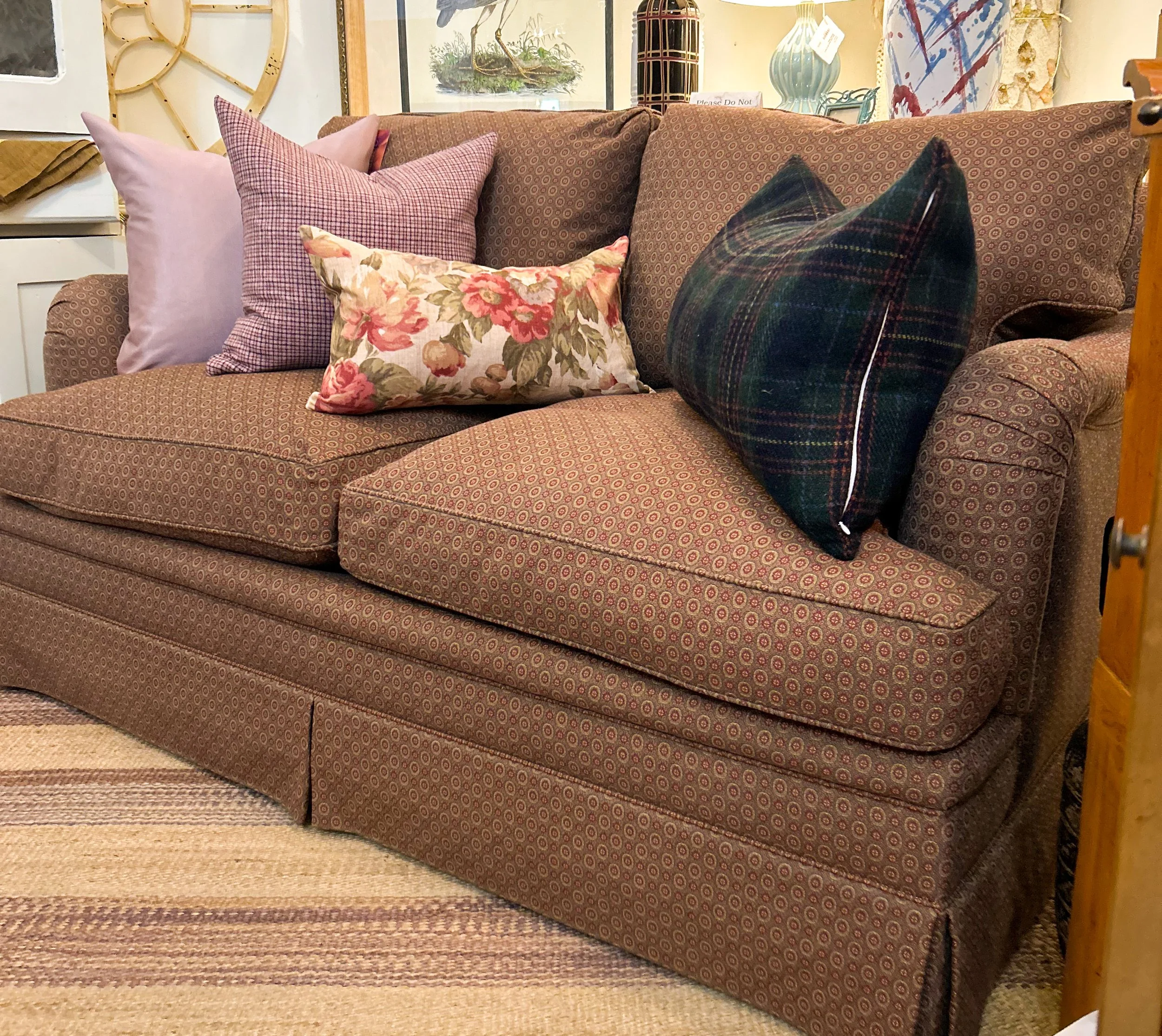 A brown patterned sofa with five decorative pillows, including one with a floral design, another with a plaid pattern, and a solid lavender pillow. The background features artwork and decorative objects.