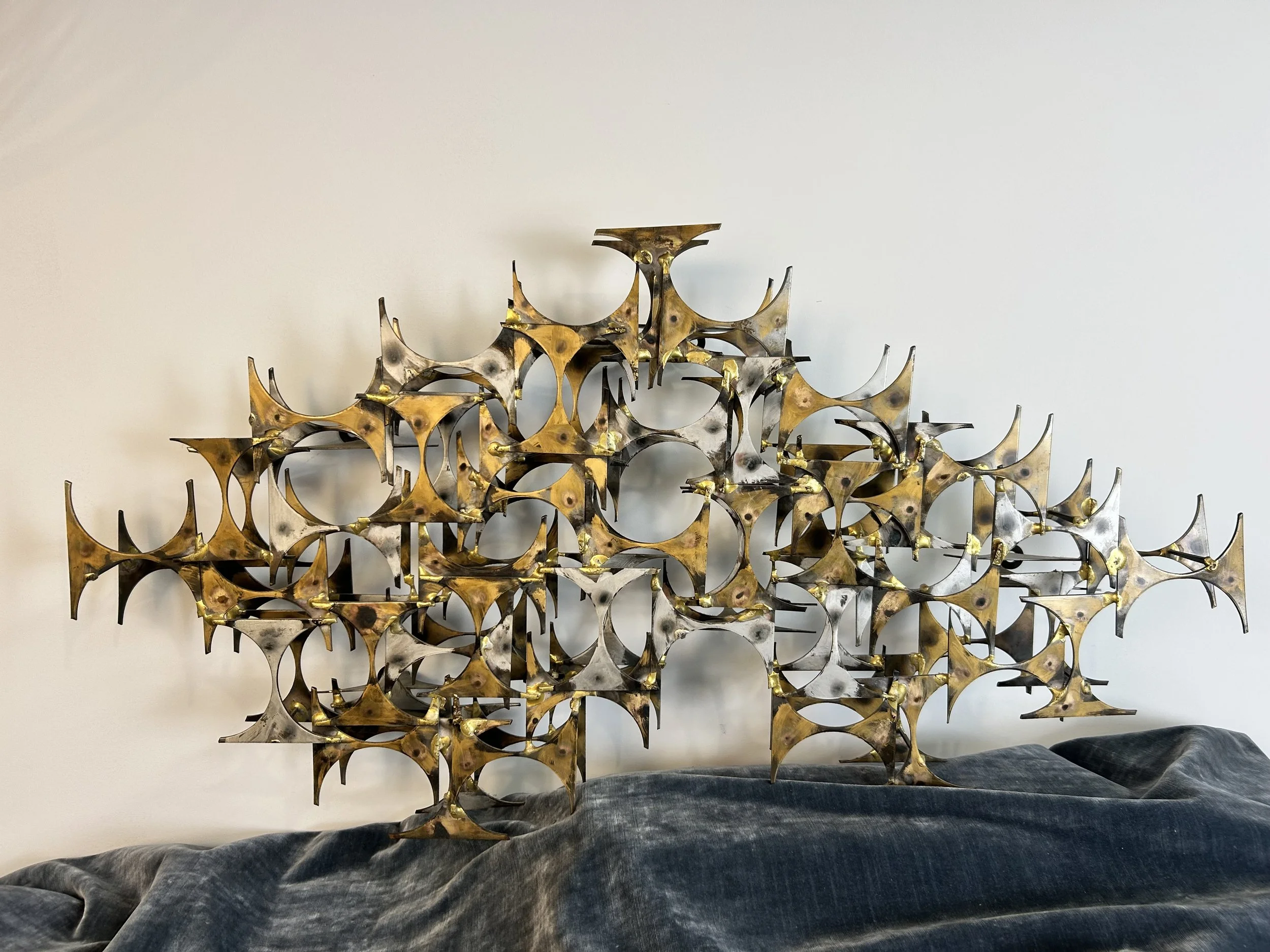 Metal wall art sculpture with interconnected crescent shapes and circular cutouts, placed on a black fabric-covered surface against a plain white wall.