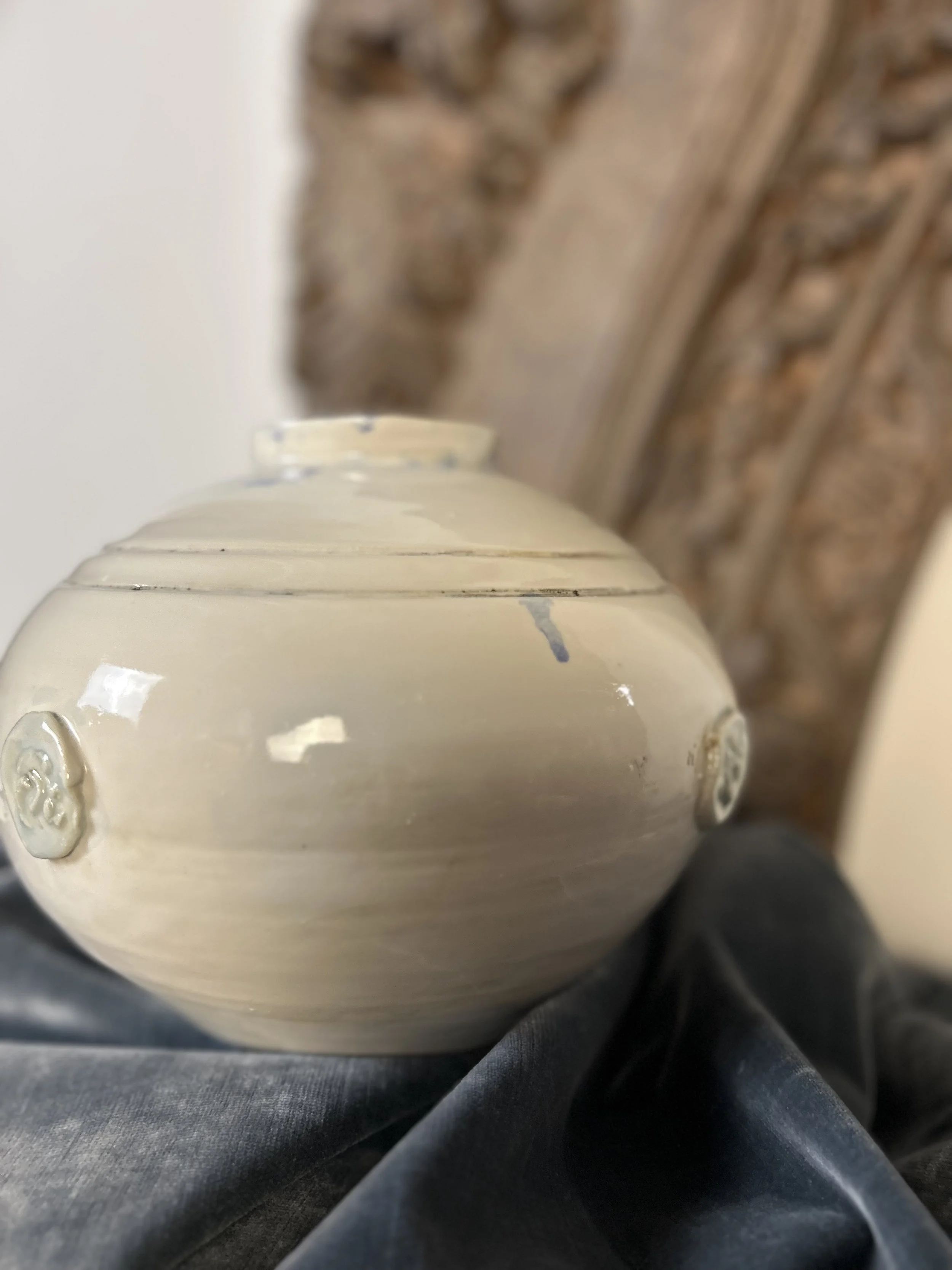 Close-up of a cream-colored ceramic vase with two decorative buttons on its side, resting on black fabric with a carved wooden frame in the background.