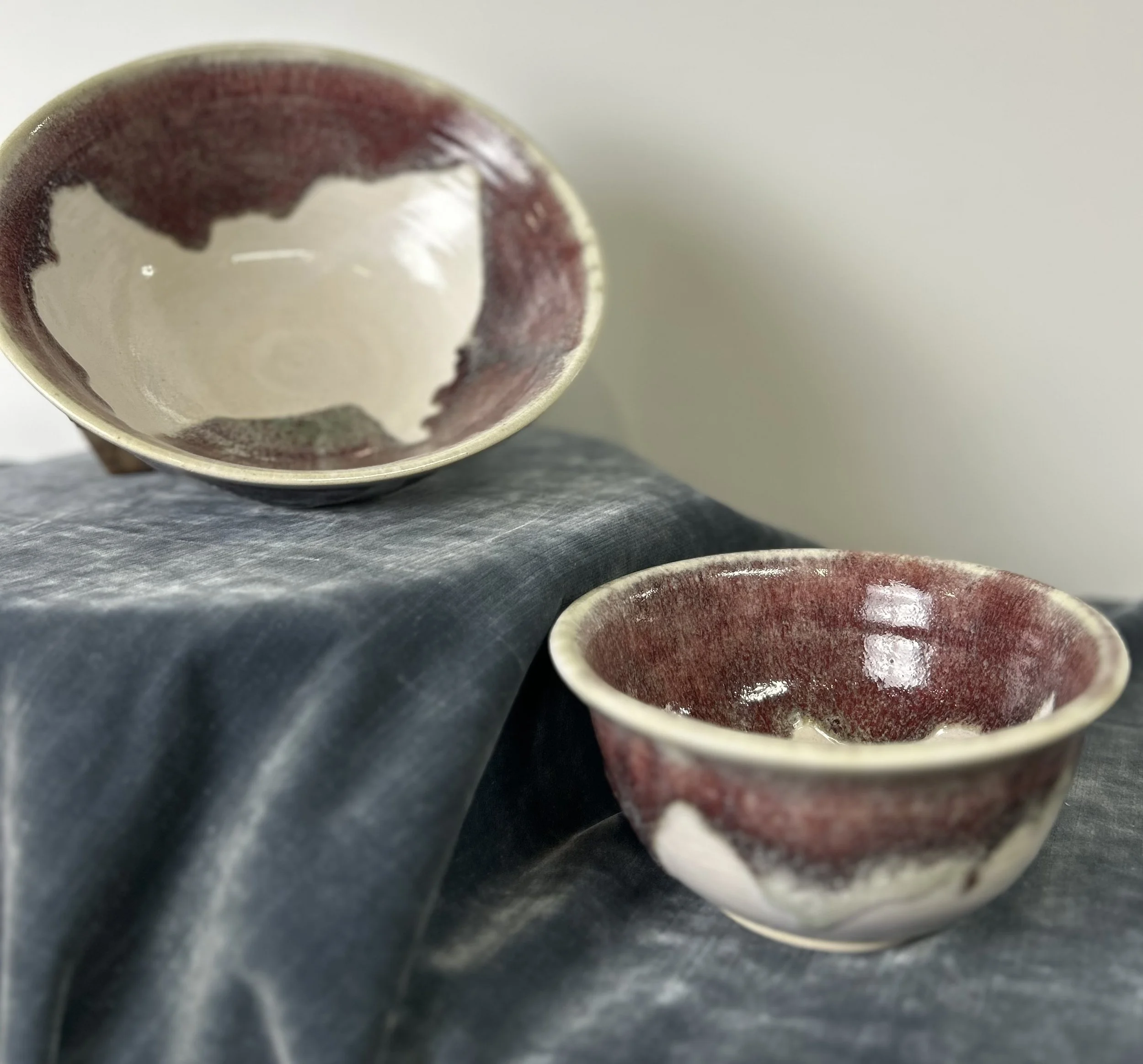 Two ceramic bowls with a maroon and cream glaze, one held upside down on a dark fabric surface and the other placed on the fabric.
