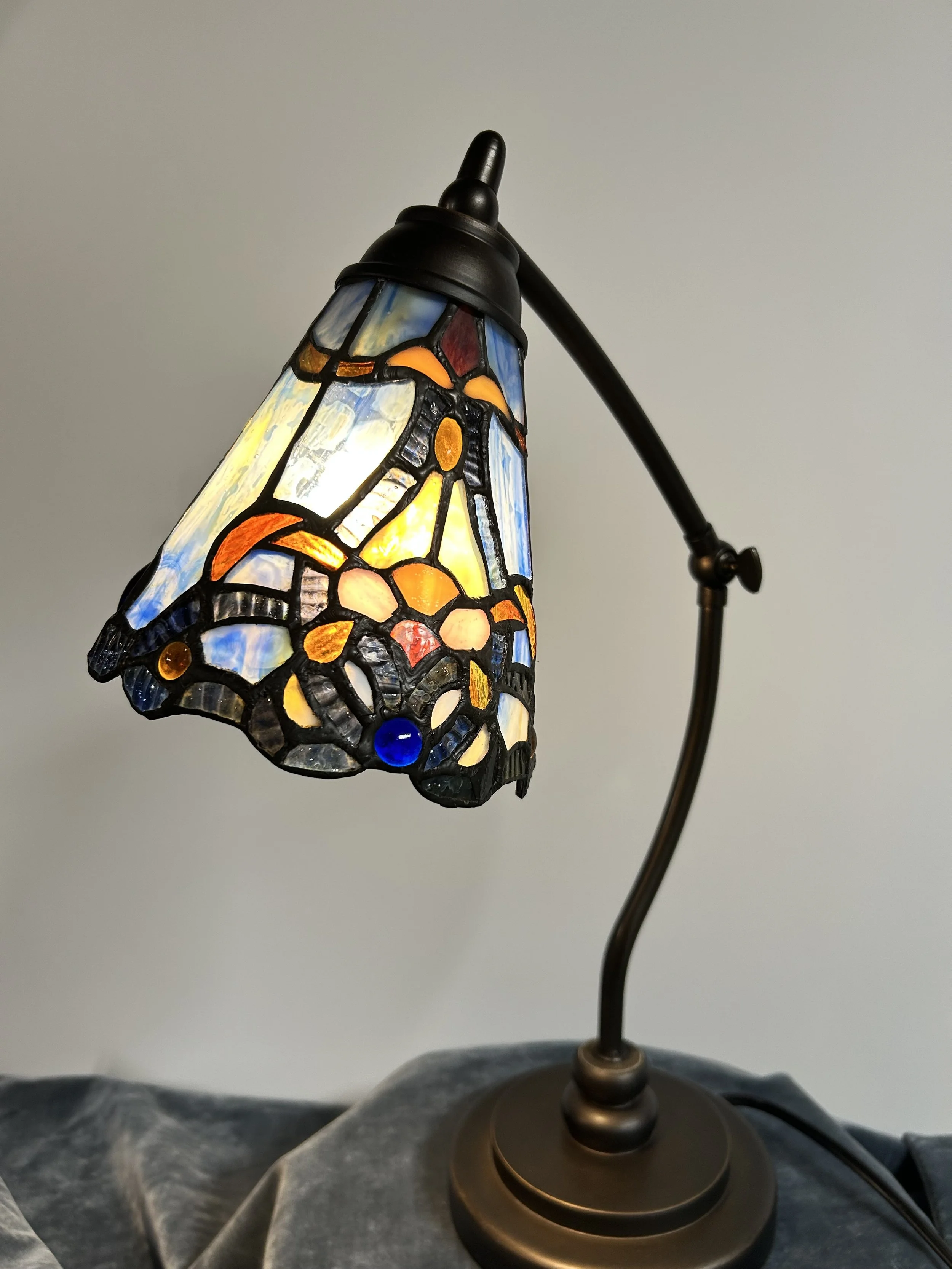 A Tiffany-style stained glass table lamp with a floral design on the shade, turned on, on a gray surface against a plain background.