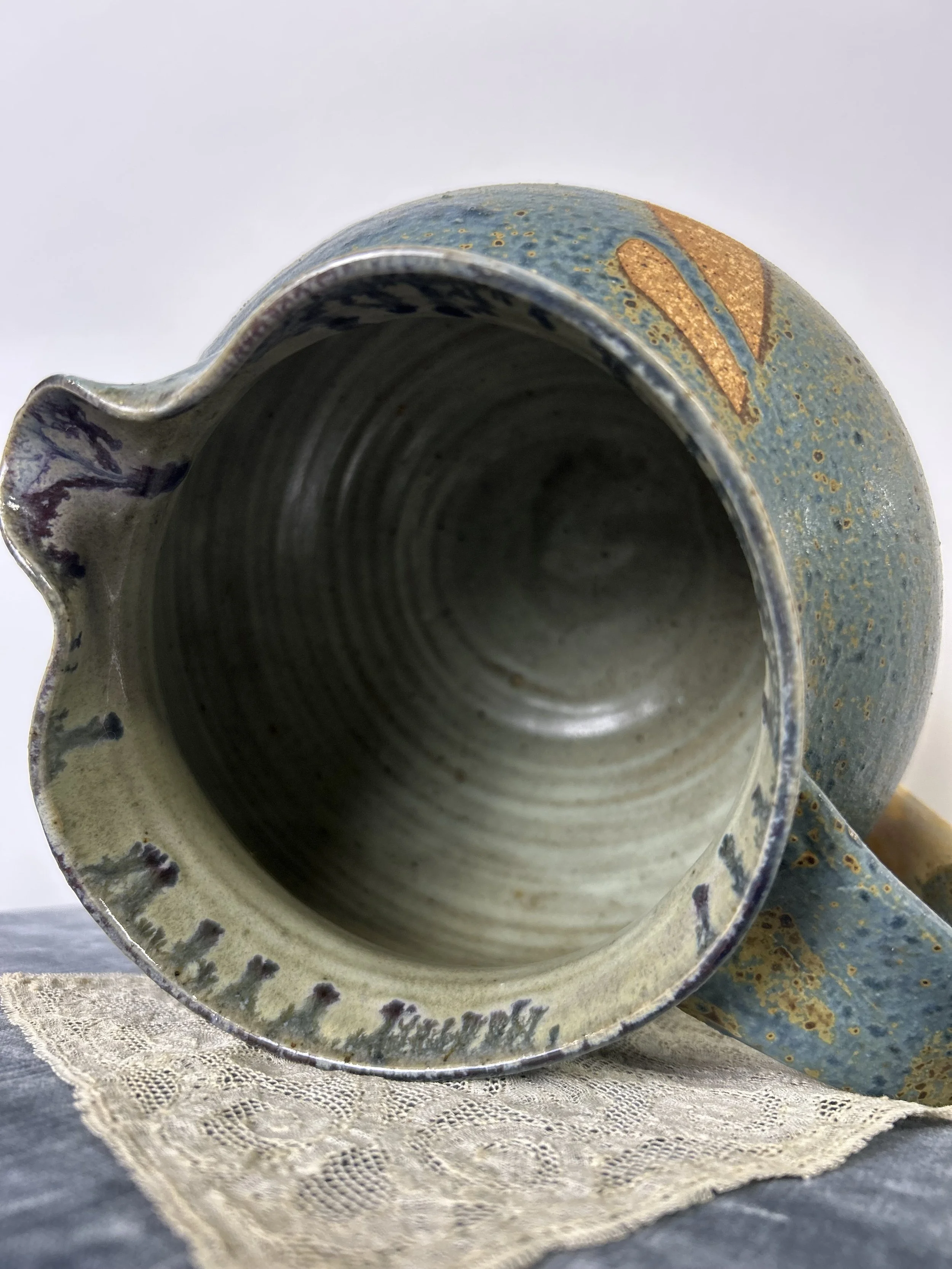 Close-up of a ceramic mug with a spout, showing the interior and exterior with blue, beige, and gold accents, placed on a lace cloth.
