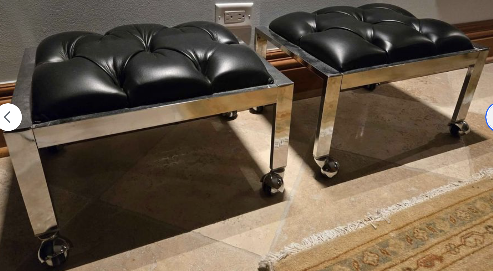 Two stools with black tufted leather cushions and chrome frames on wheels, placed on a tiled floor near a wall with an electrical outlet.