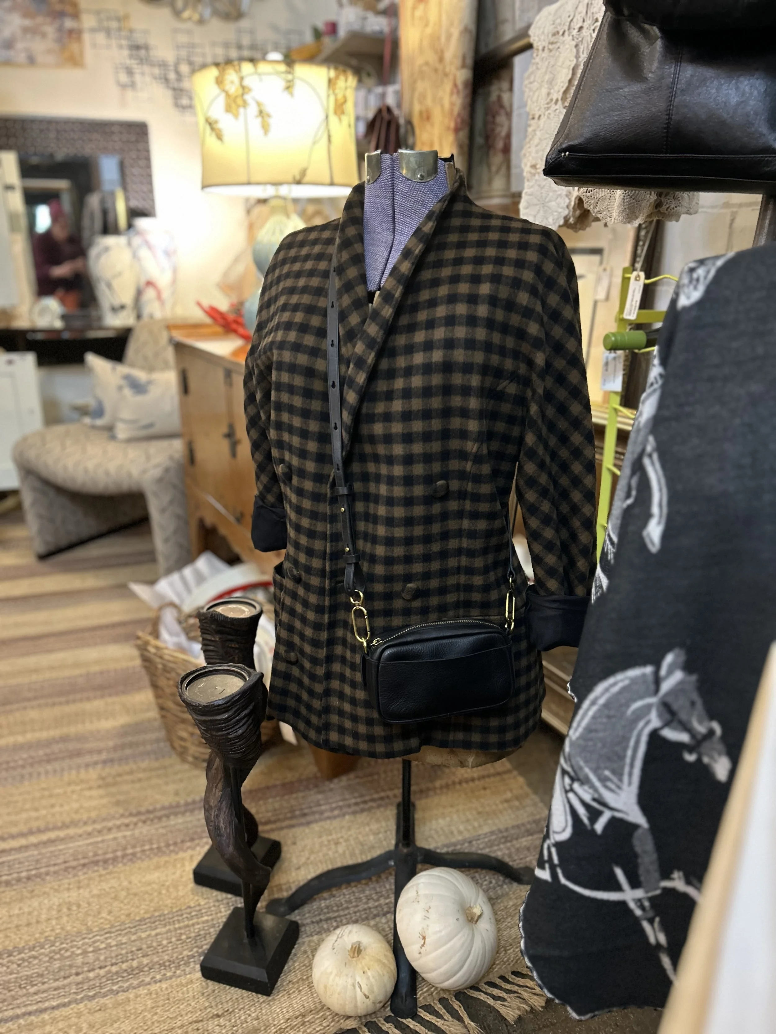 Mannequin dressed in a brown and black checkered coat, with a black leather handbag hanging from the shoulder, and a necklace. The display is set in a vintage or antique store with various home decor items and furniture around, including a lamp, pump
