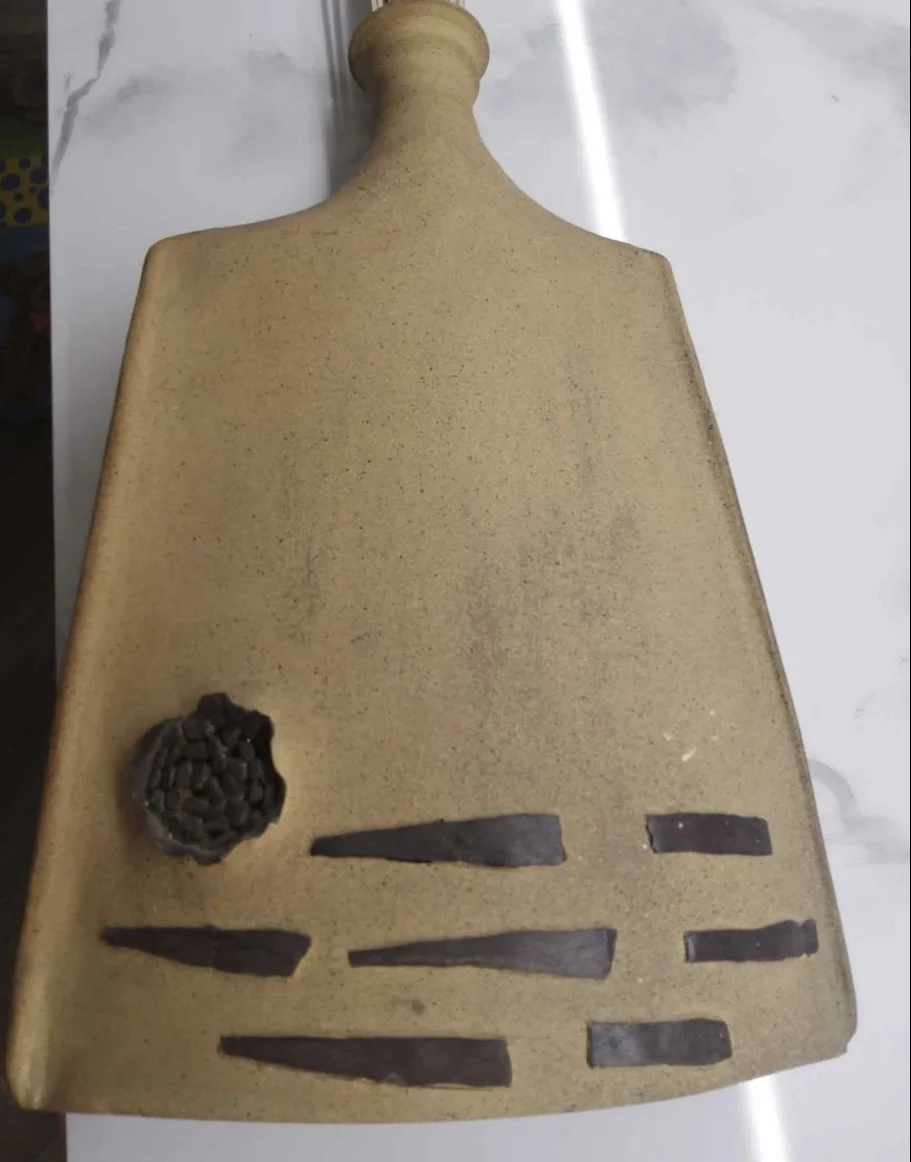 Ceramic sculpture resembling a human torso with textured surface, featuring abstract black clay or metal embellishments on the lower left and right sides.