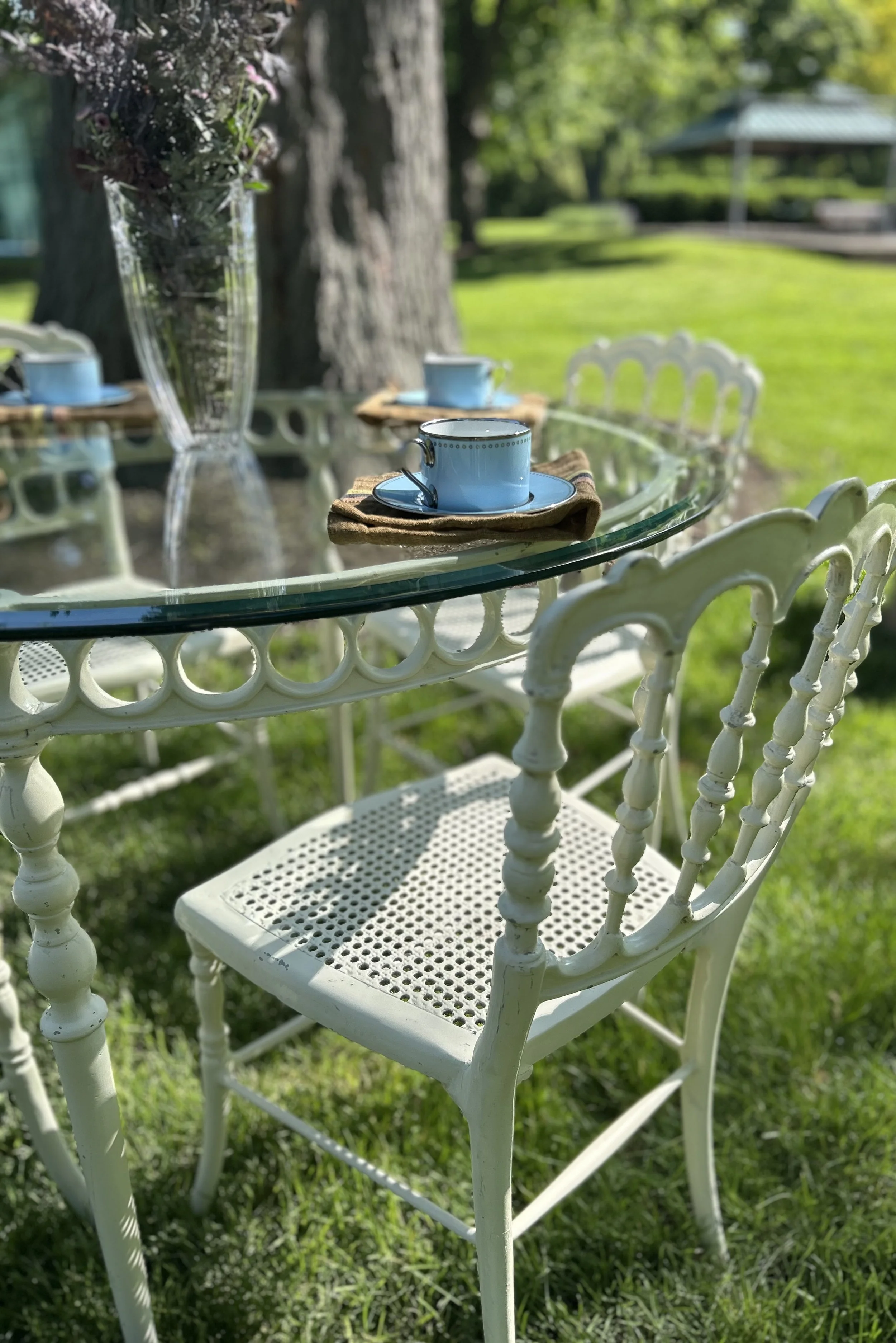 An outdoor table set with blue tea cups and saucers on beige napkins, in a garden with green grass, trees, and a gazebo in the background.