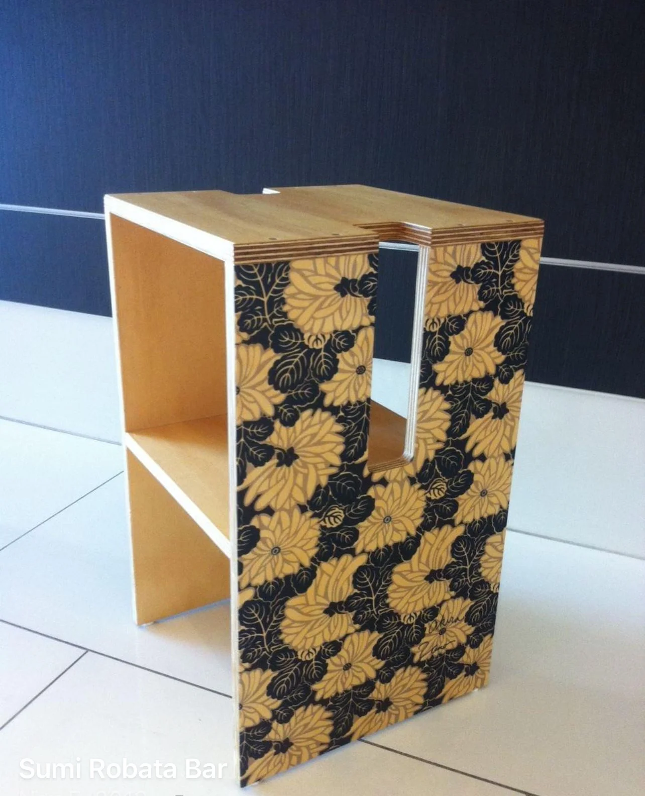 A modern chair with a rectangular cut-out in the center, featuring a floral pattern in yellow and black on the side panels, and a wooden top and shelf. The chair is placed on a tiled floor against a dark wall. Japanese artist. 