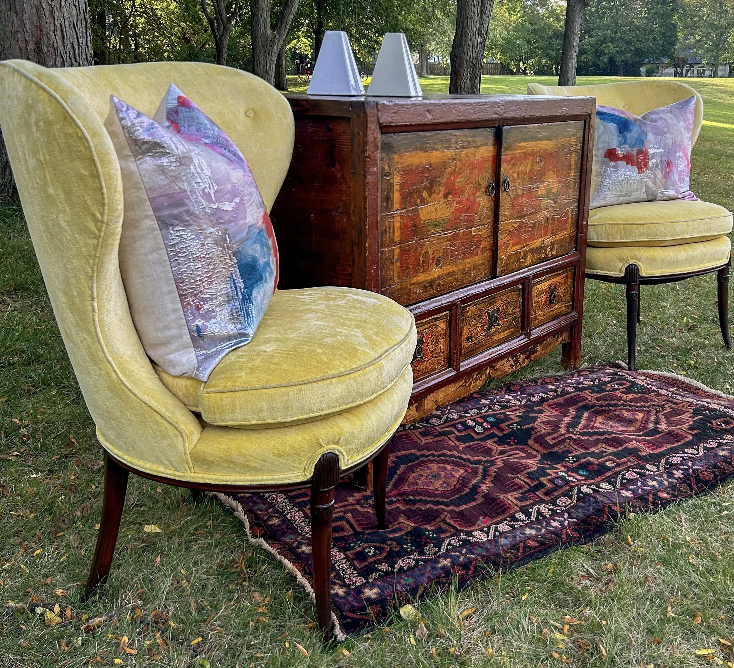 Yellow velvet armchairs with decorative pillows flanking a wooden cabinet, placed on a patterned rug outdoors on grass with trees in the background.